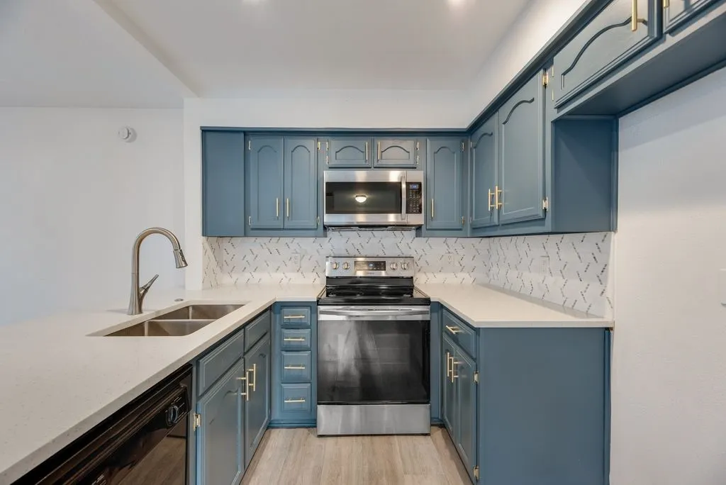 Kitchen featuring blue cabinetry with gold-toned hardware, stainless steel appliances, a double basin sink, light-toned countertops, and a decorative tile backsplash