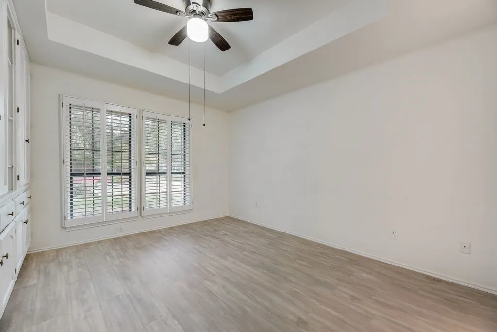 Tray ceiling with a ceiling fan, light wood-finish flooring, built-in cabinetry, and white plantation shutters on dual windows