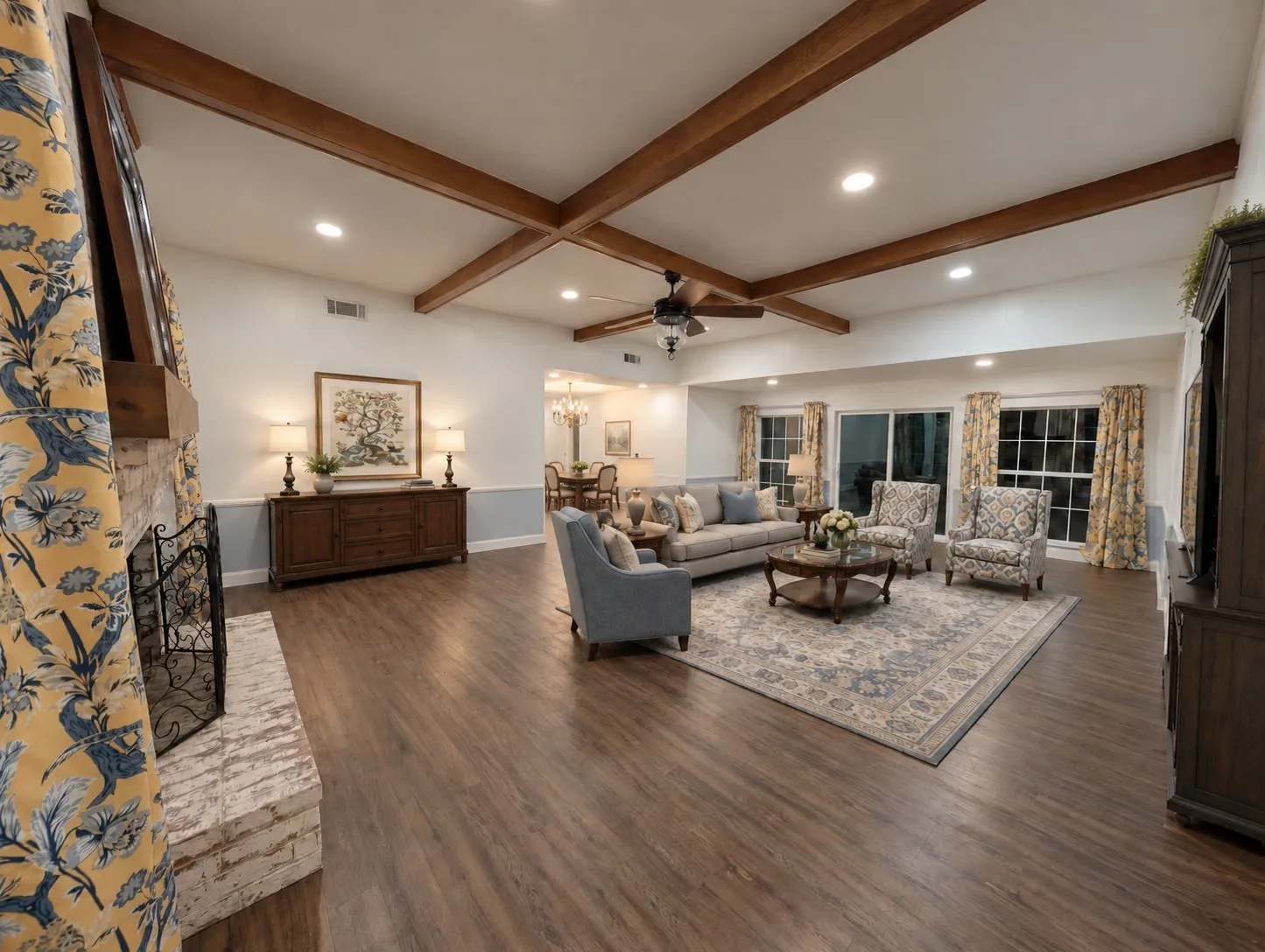 Expansive living area featuring exposed ceiling beams, wood-finish flooring, a white brick fireplace, recessed lighting, and large windows