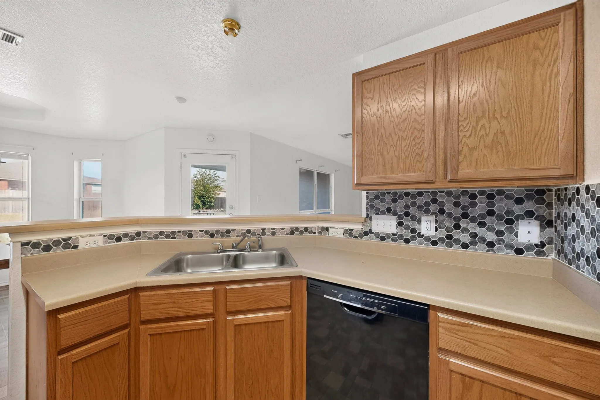 Wood-finish cabinetry, dual basin stainless steel sink, black dishwasher, and hexagonal mosaic tile backsplash
