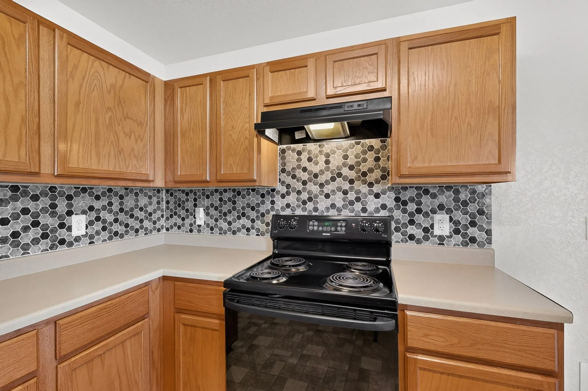 Natural wood-finish cabinetry, black electric range with hood, hexagon tile backsplash, and light-toned countertops