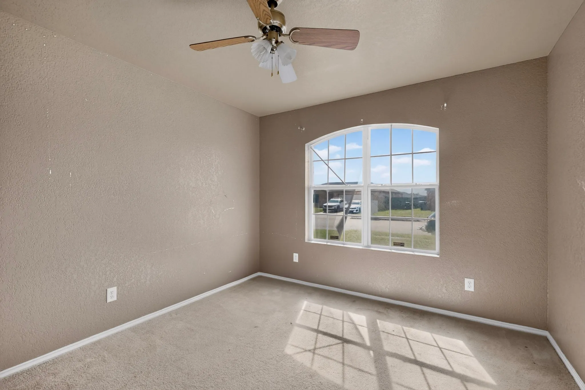 Arched window providing natural illumination, ceiling fan with light fixture, neutral-toned walls, and carpeted flooring