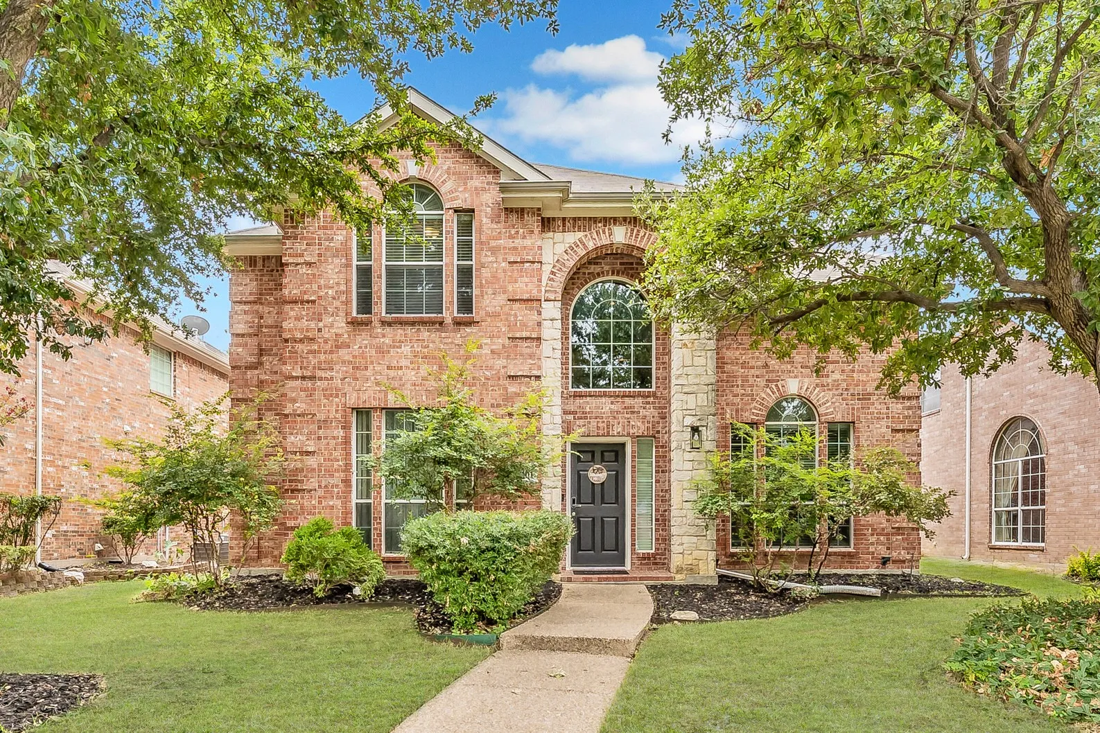 Brick and stone facade with arched windows, mature landscaping, and an inviting front walkway