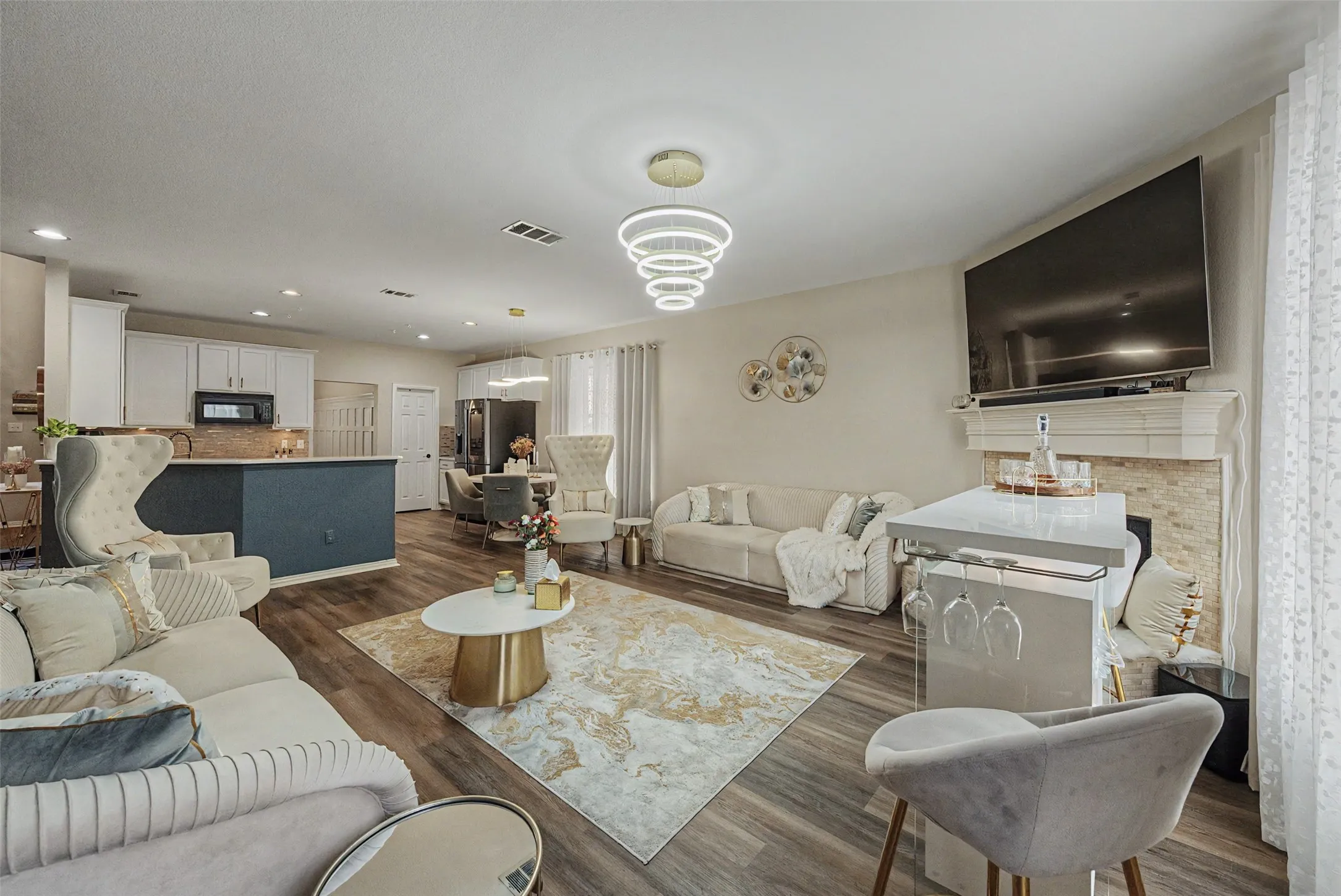 Open-concept living space featuring wood-finish flooring, a contemporary tiered chandelier, white kitchen cabinetry with a dark island, and a fireplace with a stone surround and mantel