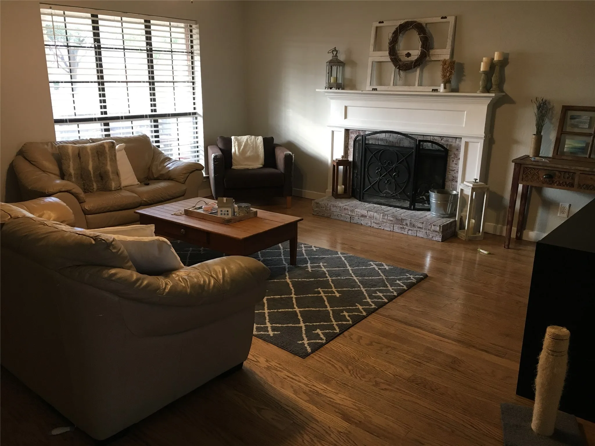 Hardwood flooring throughout, a large window with horizontal blinds, a fireplace with a brick surround, and a white mantel