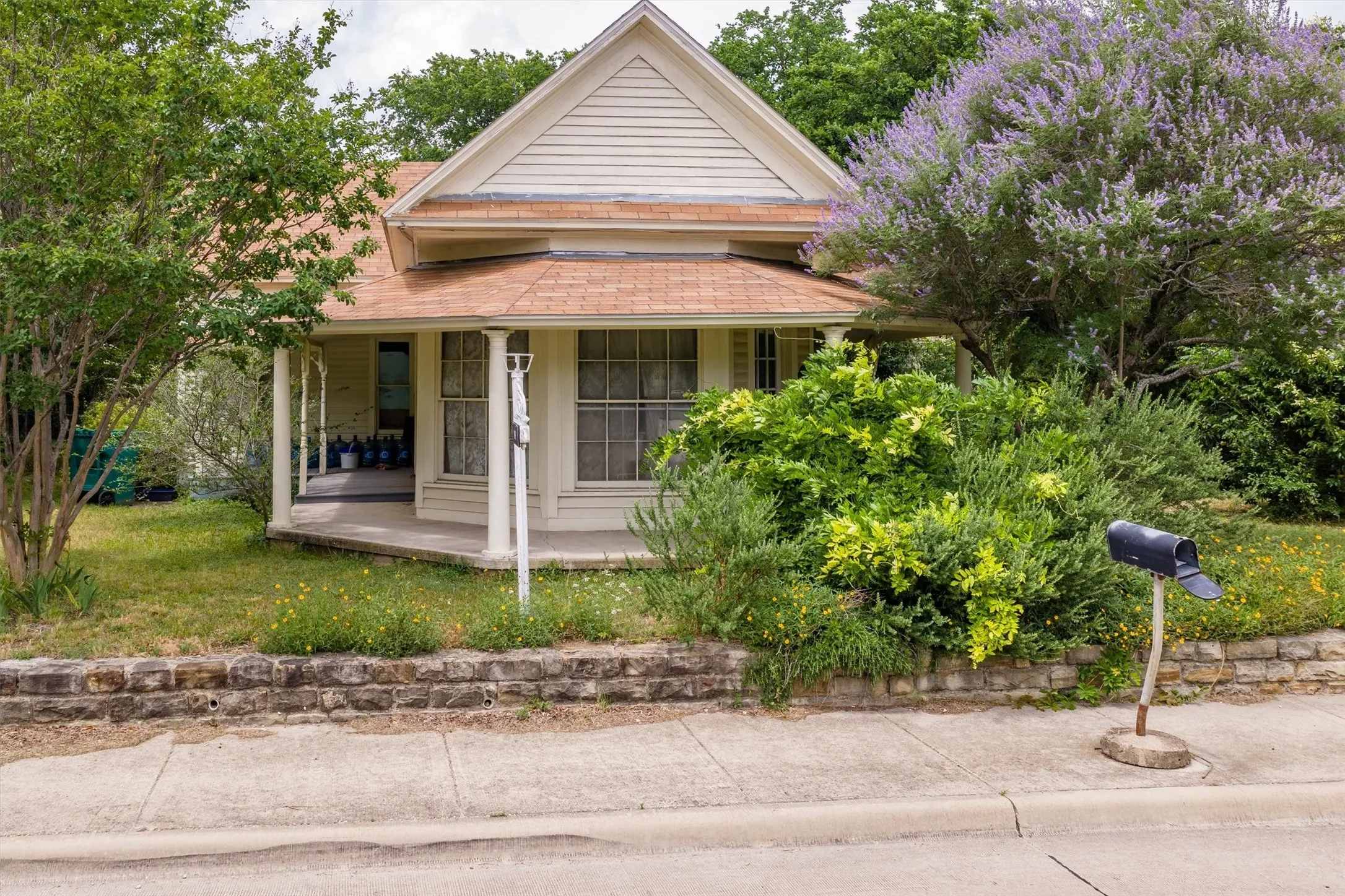 Gable roofline, covered front porch with pillars, prominent front windows, mature landscaping, and a stone retaining wall