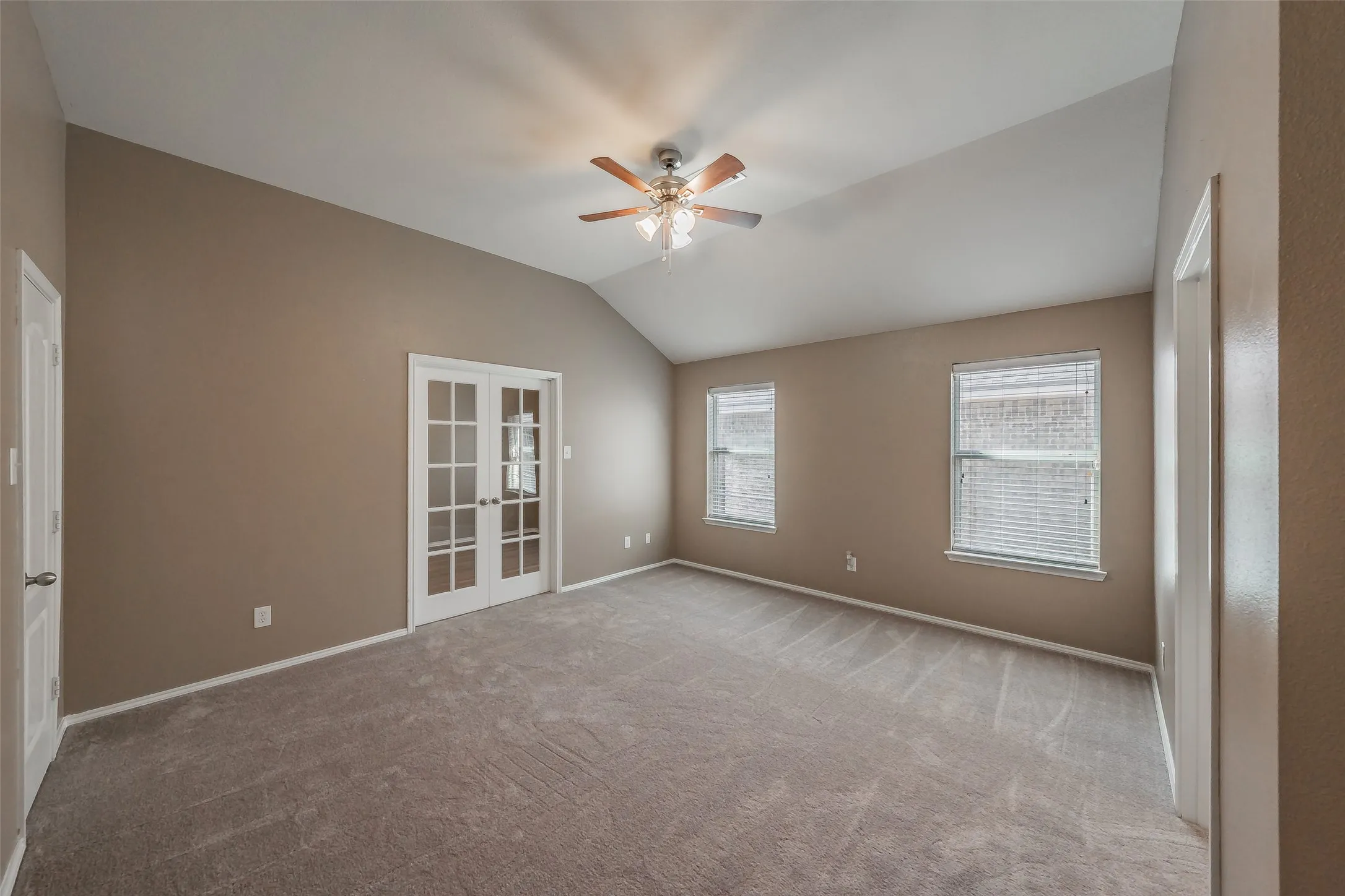 Vaulted ceiling, two windows with blinds, French doors, ceiling fan with light, and light-toned carpet