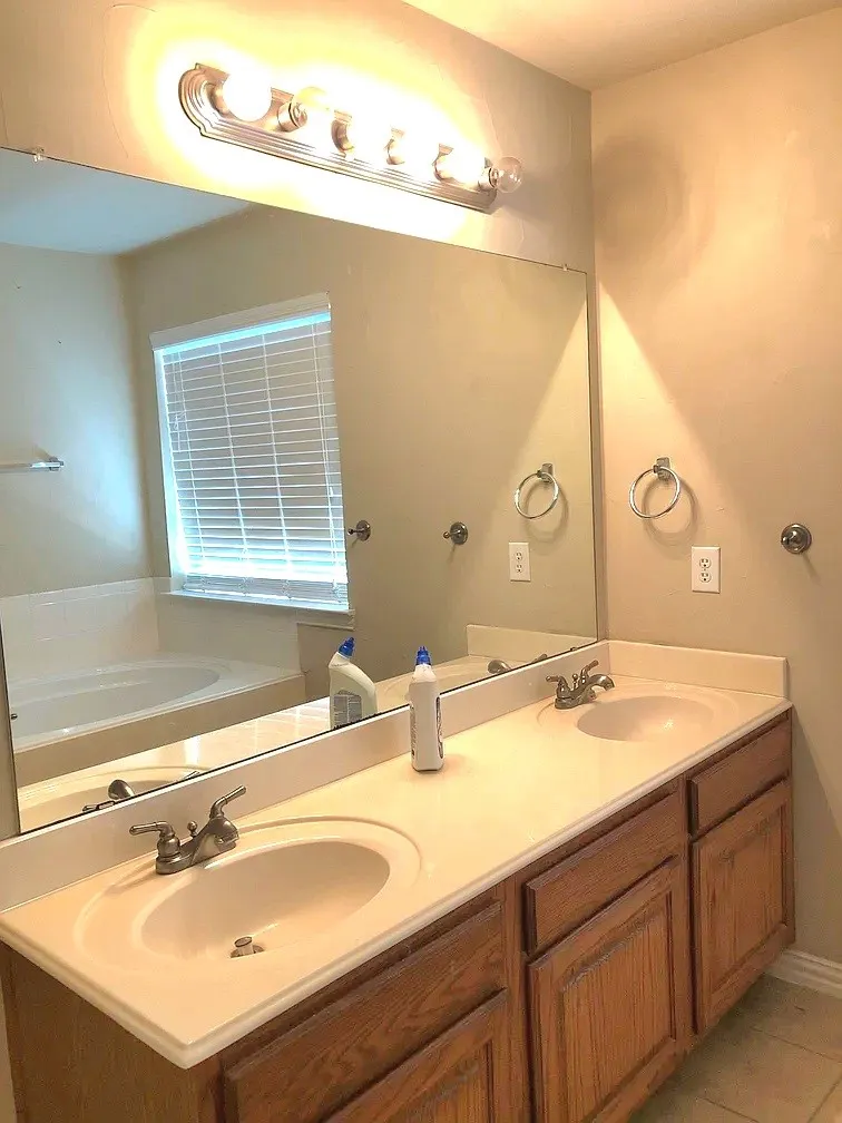 Dual vanity with integrated sinks, wood-finish cabinetry, a large wall-mounted mirror, and a built-in tub with a window