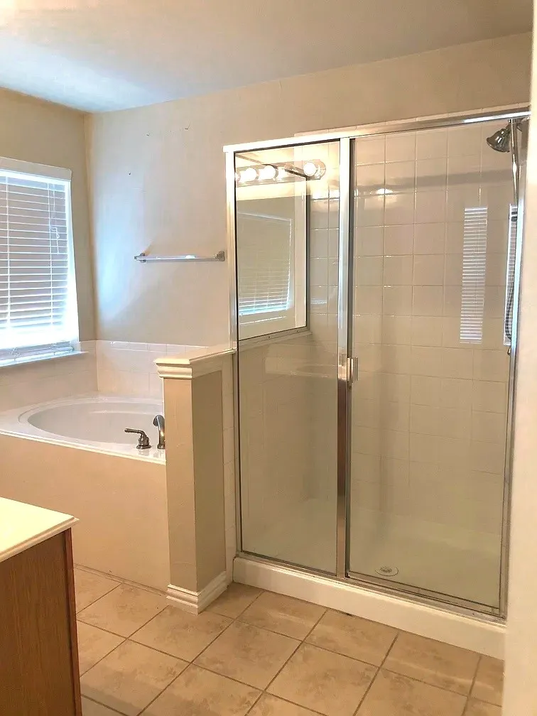 Tile flooring, built-in bathtub, glass-enclosed shower, wall-mounted light fixture, and window with blinds