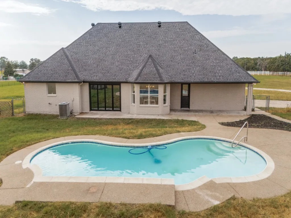 Rear exterior with a freeform swimming pool, concrete patio, sliding glass doors, a bay window, and a dark shingle roof