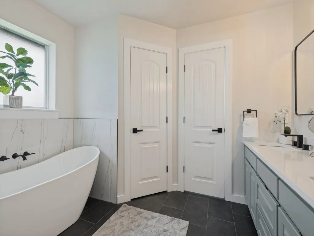 Freestanding soaking tub with wall-mounted matte black fixtures, white stone tile wainscoting, dark tile flooring, and a double vanity with a light gray cabinet and white countertop