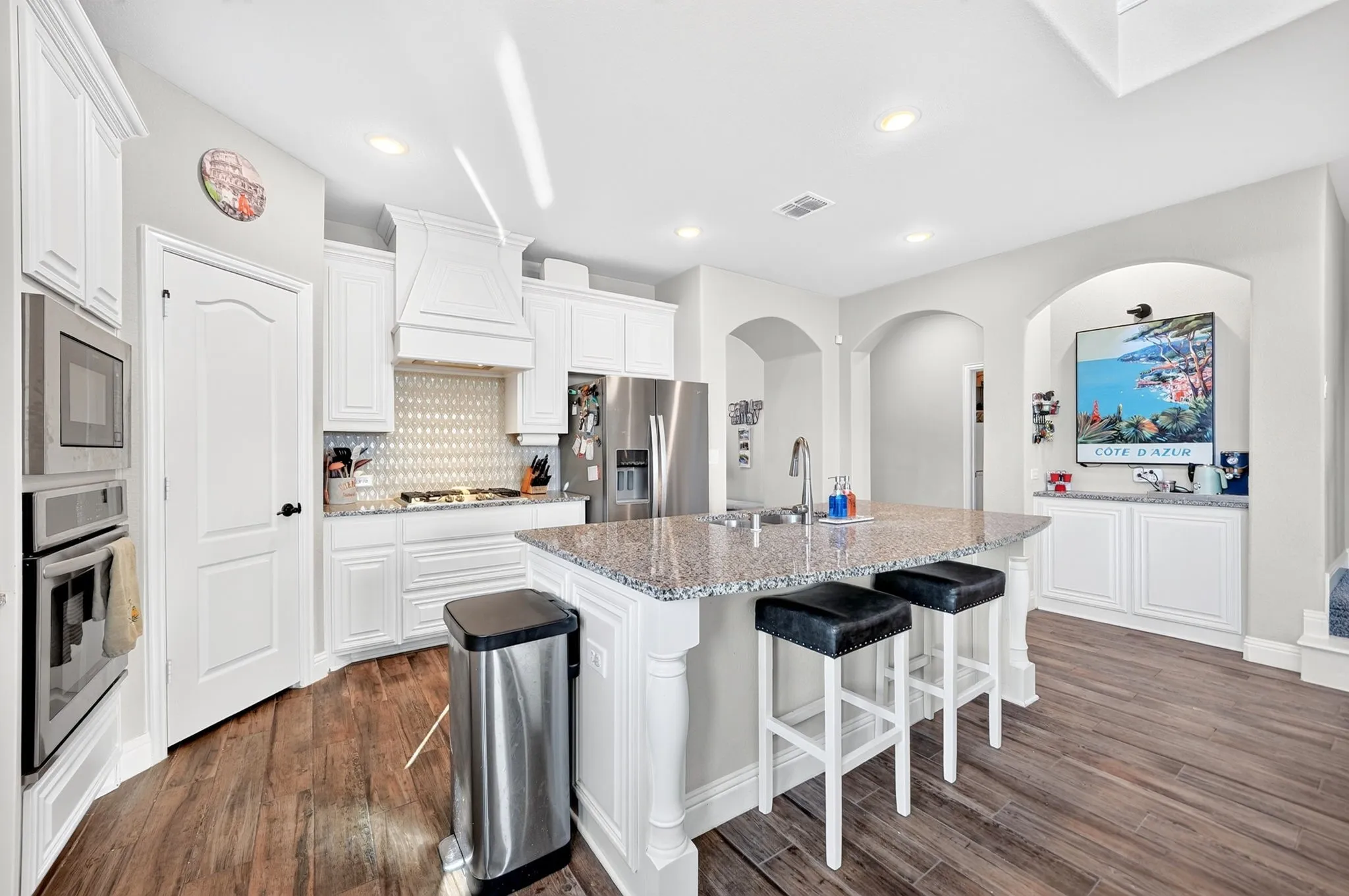Kitchen featuring wood-finish flooring, white cabinetry, a built-in oven and microwave, a center island with a sink, and recessed lighting