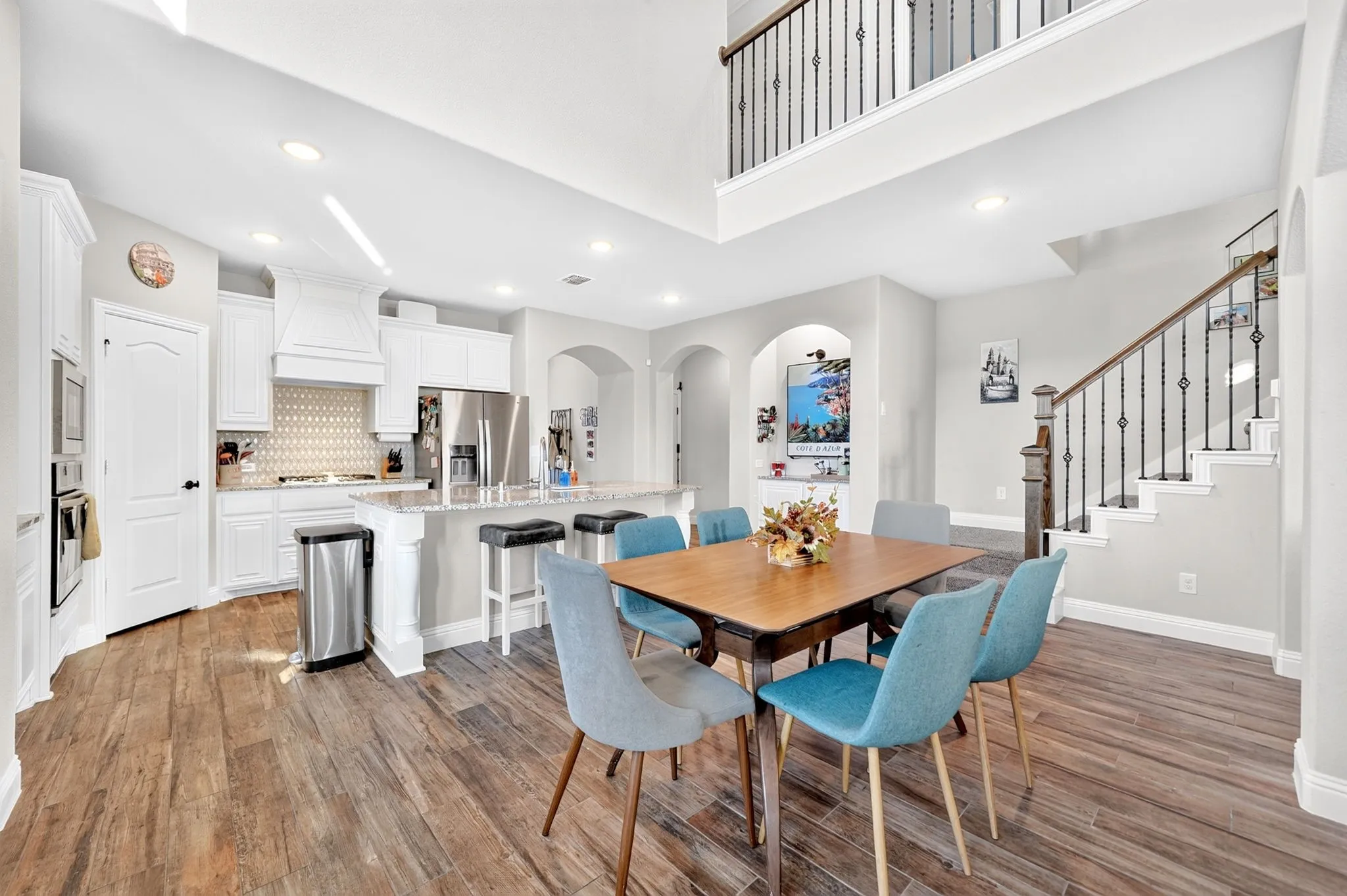 Open-concept living space featuring wood-finish flooring, a kitchen with white cabinetry and stainless steel appliances, a kitchen island with seating, a dining area, and a staircase with an ornamental iron railing