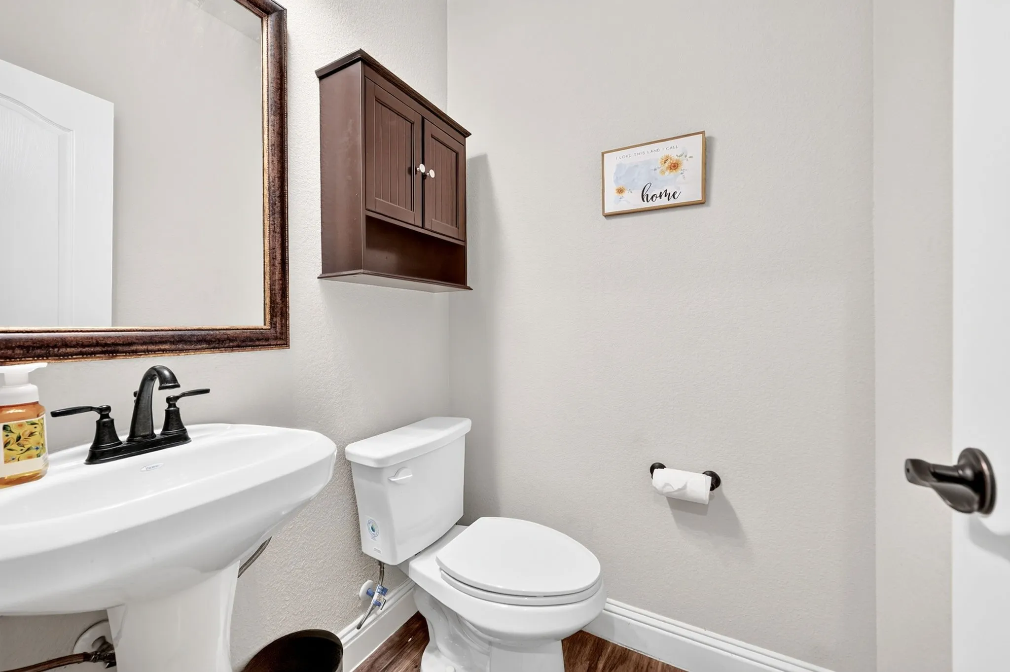 Pedestal sink with a dark bronze faucet, framed mirror, wall-mounted storage cabinet, and wood-finish flooring