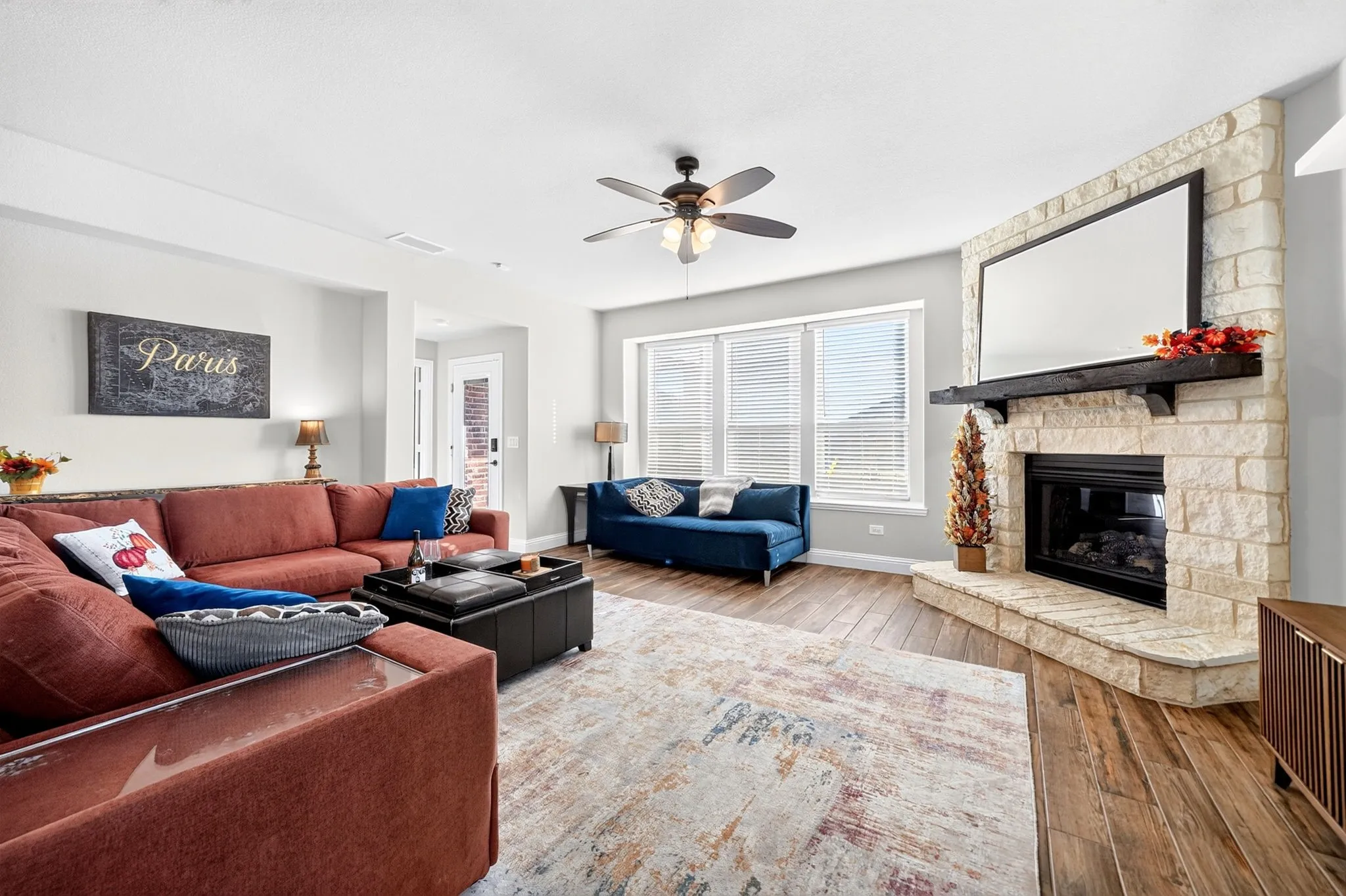 Spacious living area featuring a stone fireplace, wood-finish flooring, multiple windows with blinds, light-toned walls, and a ceiling fan