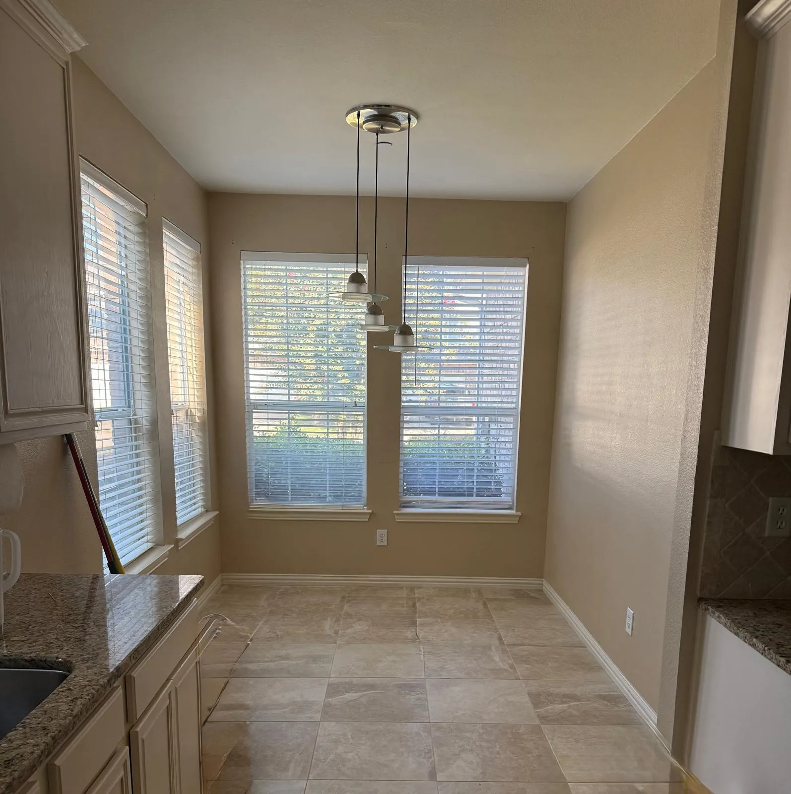 Recessed dining nook featuring ceramic tile flooring, three large windows with blinds, a multi-light pendant fixture, and a granite-topped counter with an undermount sink