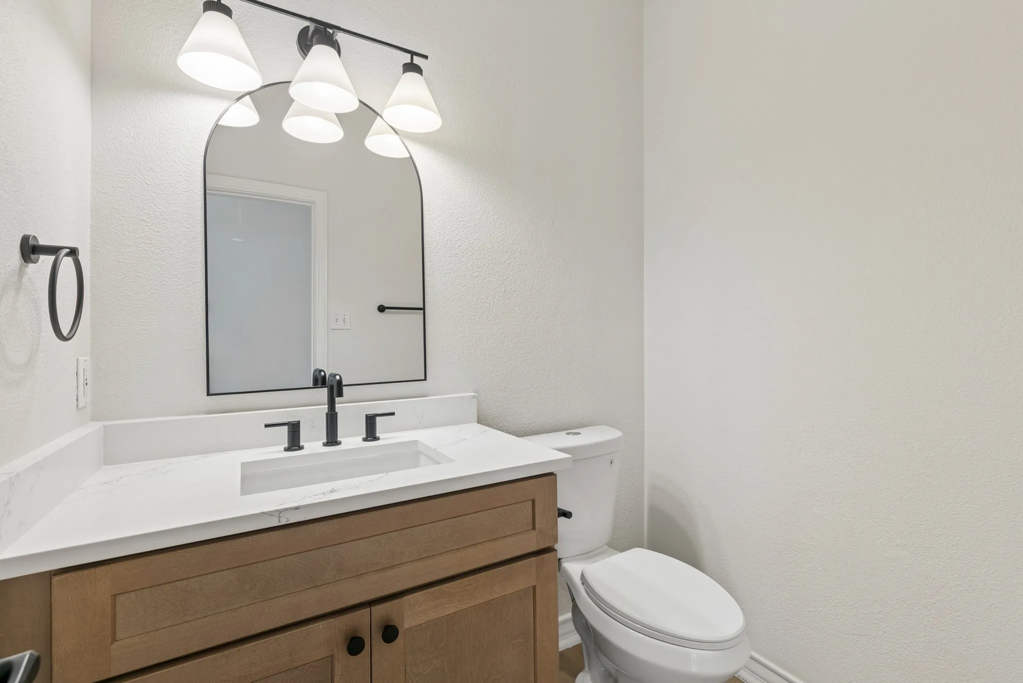 Wood-finish vanity with a white countertop and integrated sink, black hardware, arched mirror, and multi-bulb light fixture