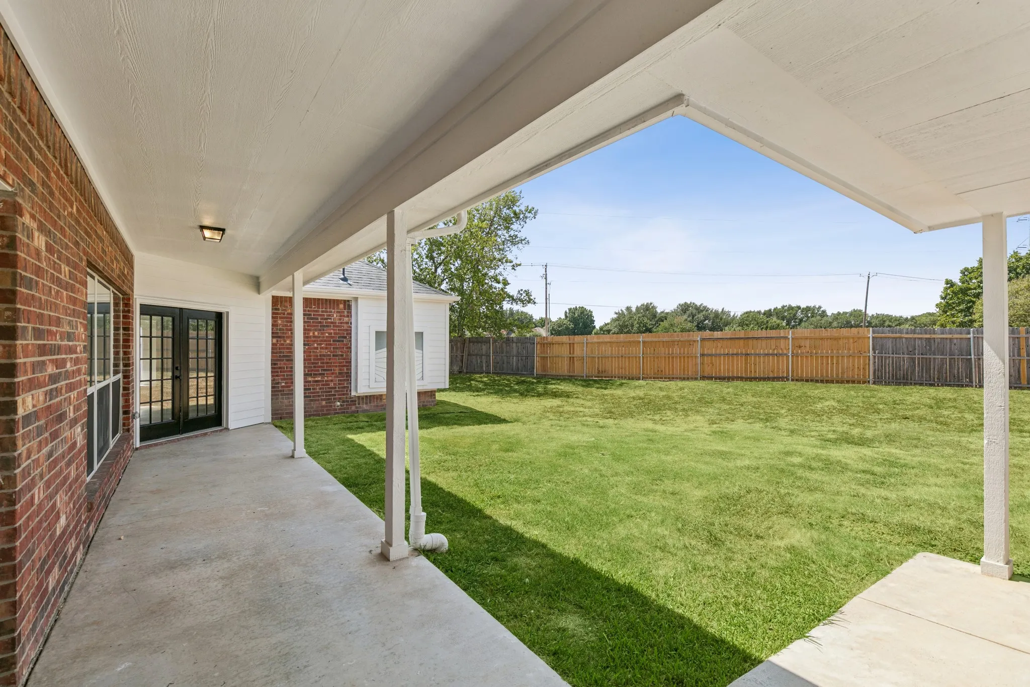Covered concrete patio with white support columns, brick exterior, French doors, and a spacious grassy yard featuring a wood fence