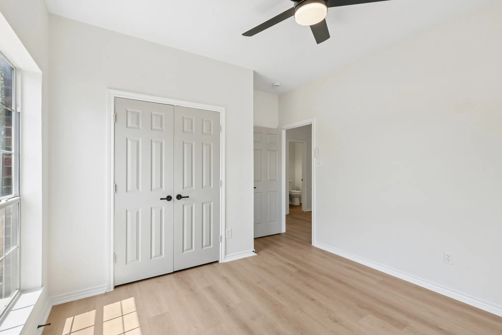 Light wood-finish flooring, white walls, and a ceiling fan with integrated lighting