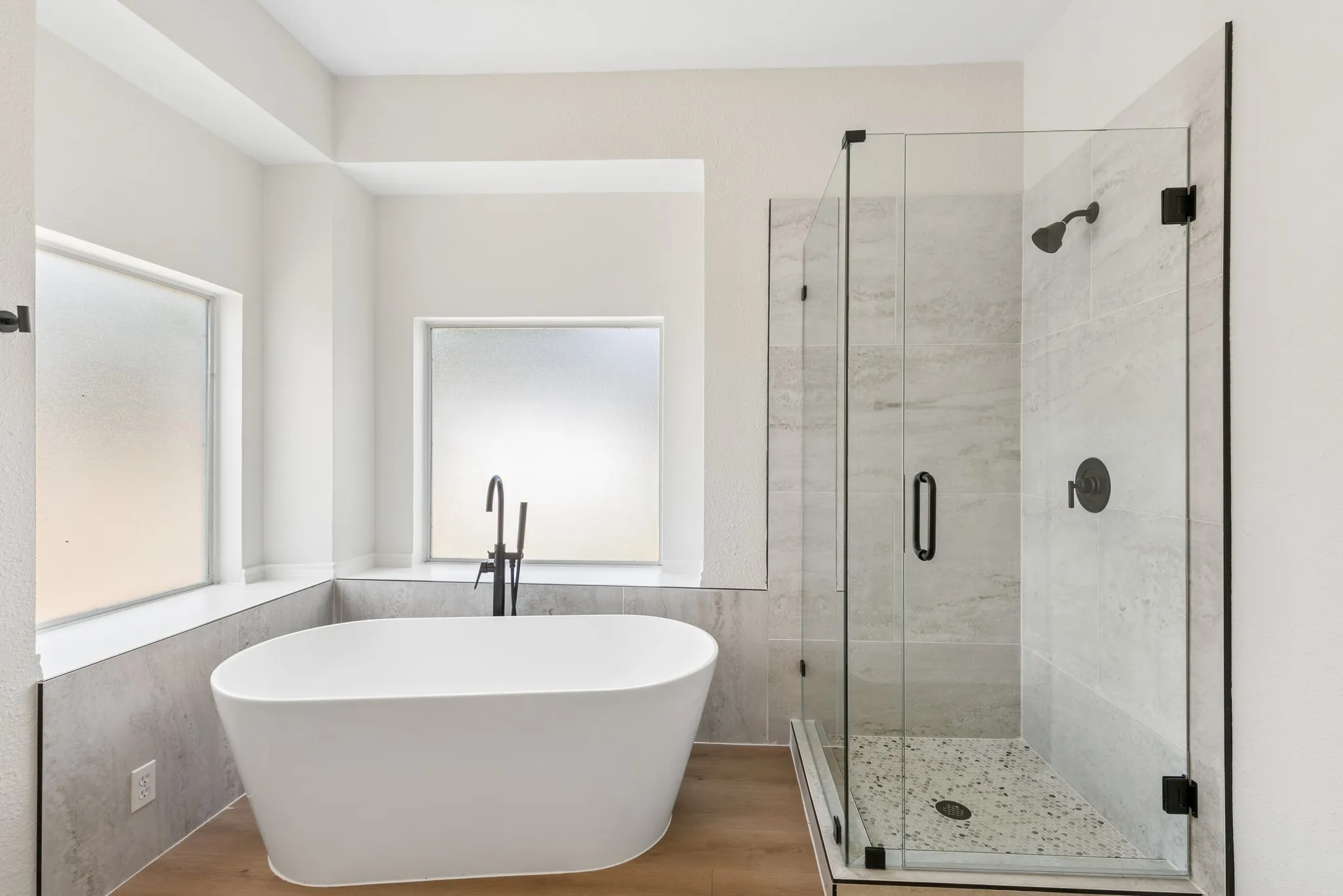 Freestanding soaking tub with matte black floor-mounted faucet, adjacent to a glass-enclosed shower with a tiled surround, and wood-finish flooring