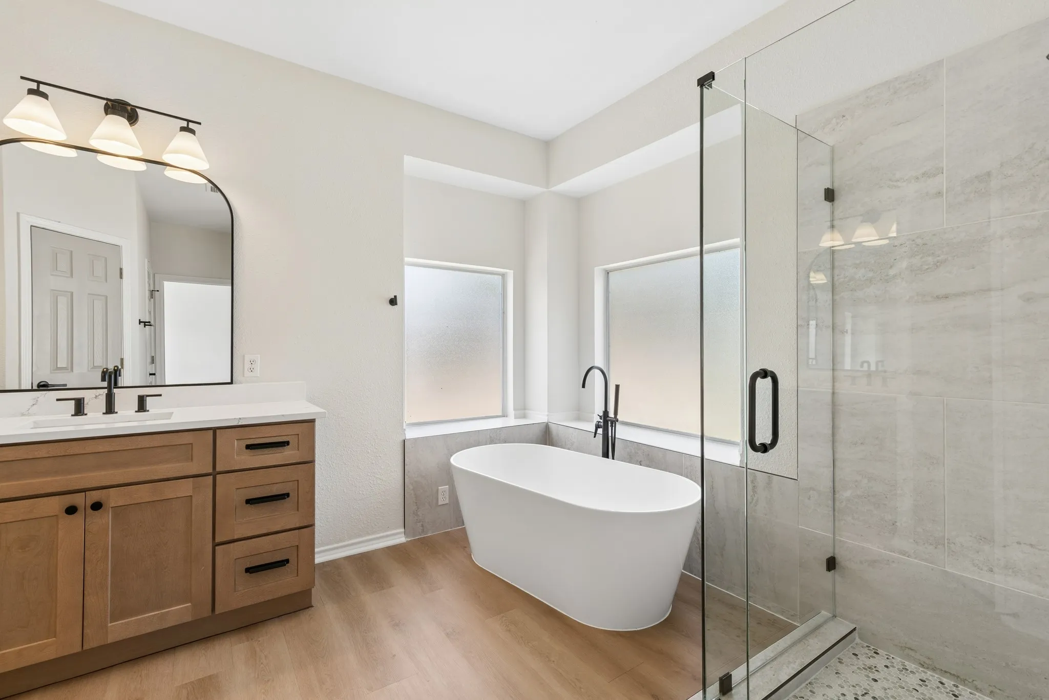 Freestanding soaking tub, frameless glass shower enclosure, wood-finish flooring, wood vanity with a white countertop, and arched mirror