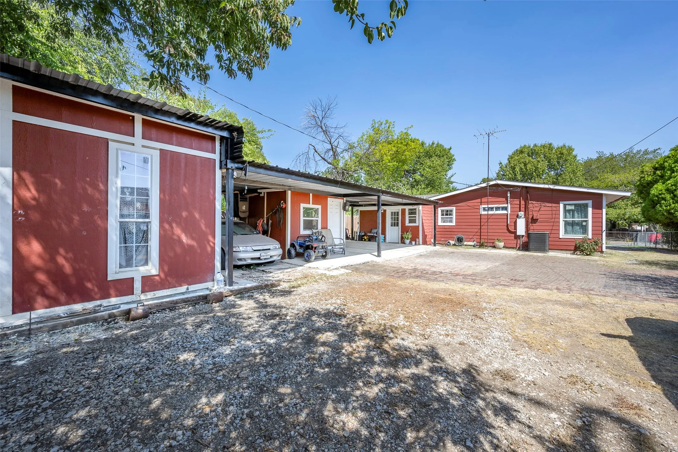 Red exterior siding, white trim windows, covered carport, gravel driveway, and a separate structure with red siding