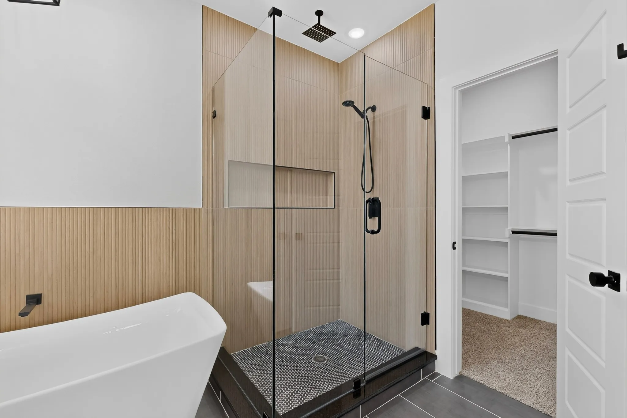Freestanding soaking tub, glass-enclosed shower with matte black fixtures, integrated shower niche, and custom wood-finish wall paneling