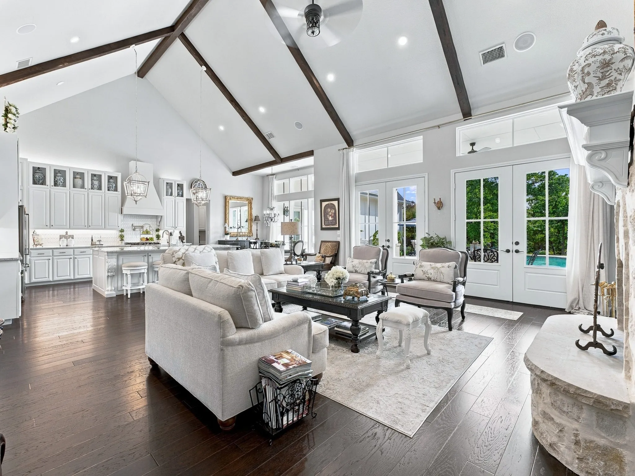 Vaulted ceiling with exposed wood beams, wood-finish flooring, built-in kitchen cabinetry, stone-clad fireplace, and double French doors