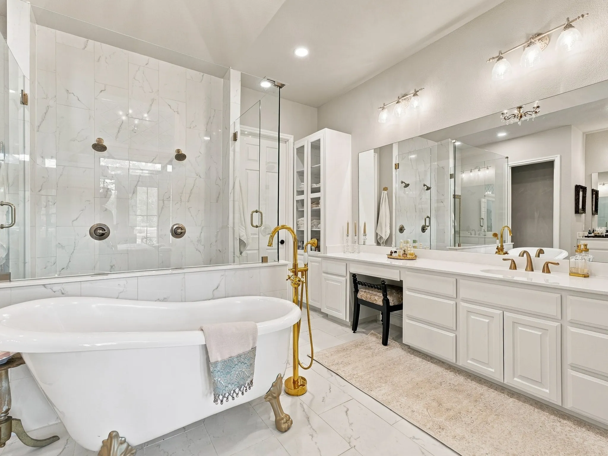 Freestanding bathtub with gold-finish fixtures, a spacious walk-in shower with multiple showerheads, dual vanities with expansive counter space, and integrated storage cabinetry