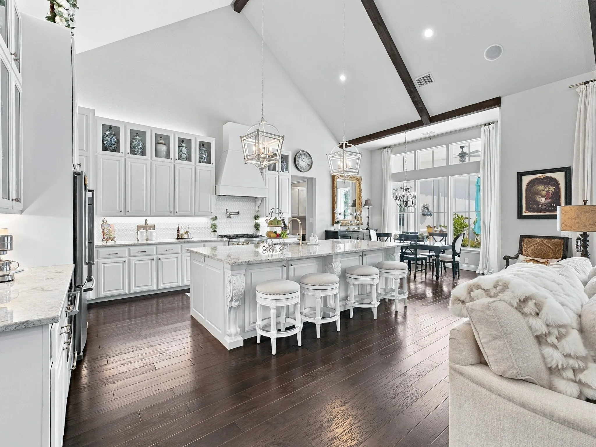 Vaulted ceiling with exposed wood beams, wood-finish flooring, white cabinetry with upper display cabinets, kitchen island with seating, and large windows with transom panels