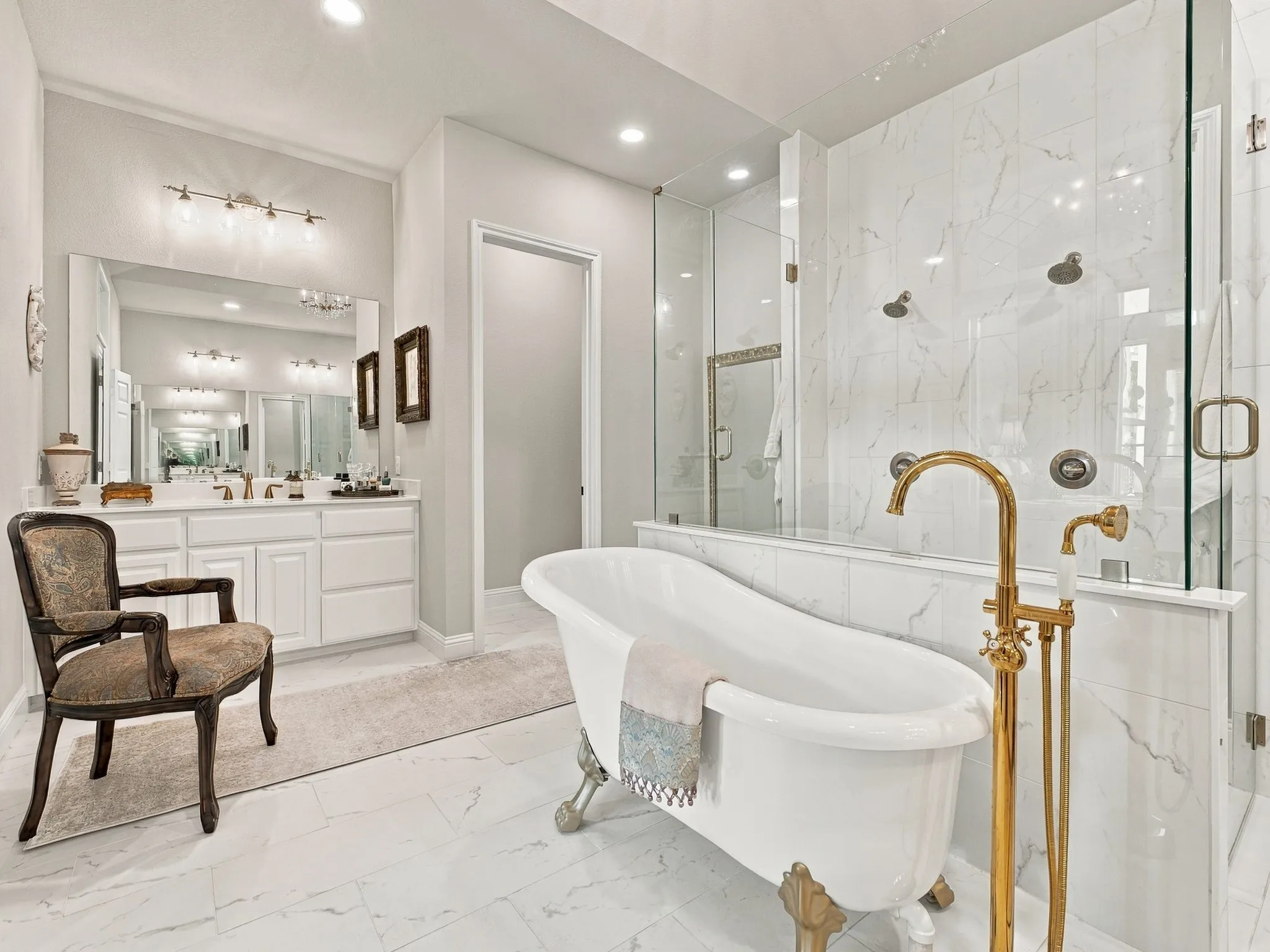 Freestanding tub with brass-finish floor-mounted faucet, dual shower system with glass enclosure, white marble-look tile, and dual vanity with white cabinetry