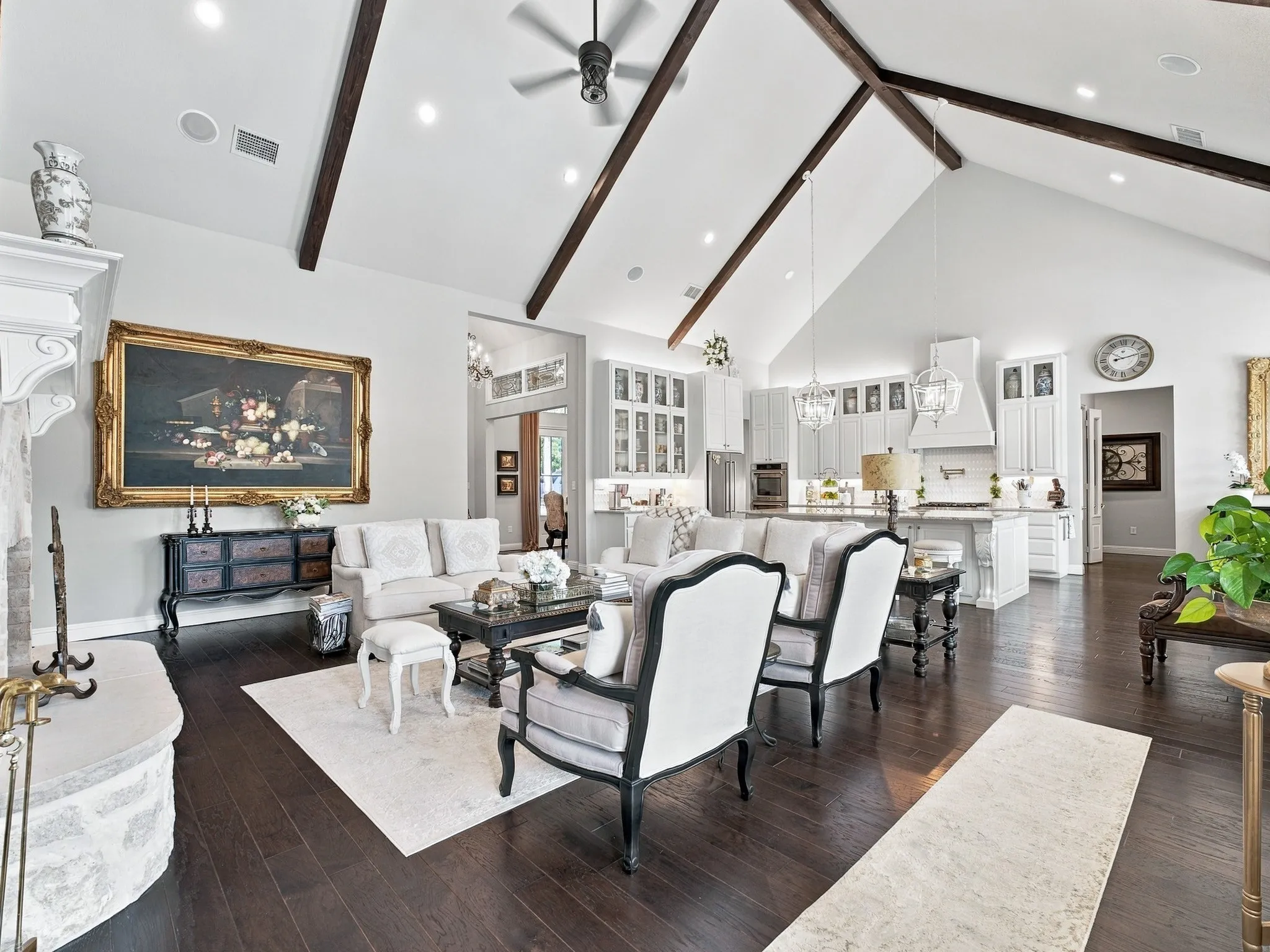 Vaulted ceiling with exposed wood beams, stone fireplace, wood-finish flooring, recessed lighting, and a kitchen featuring white cabinetry and an island