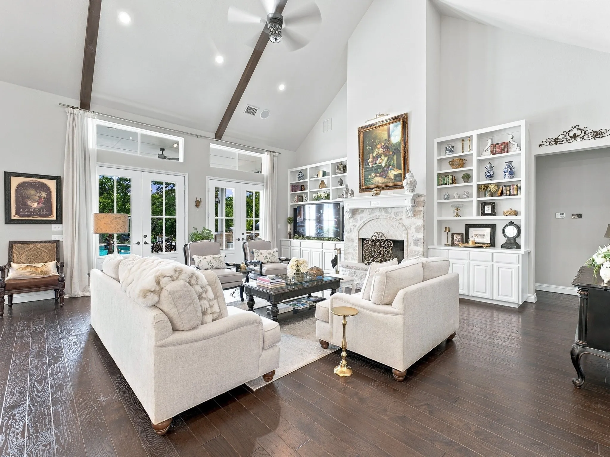 Vaulted ceiling with exposed wood beams, wood-finish flooring, built-in shelving, stone-clad fireplace, and French doors