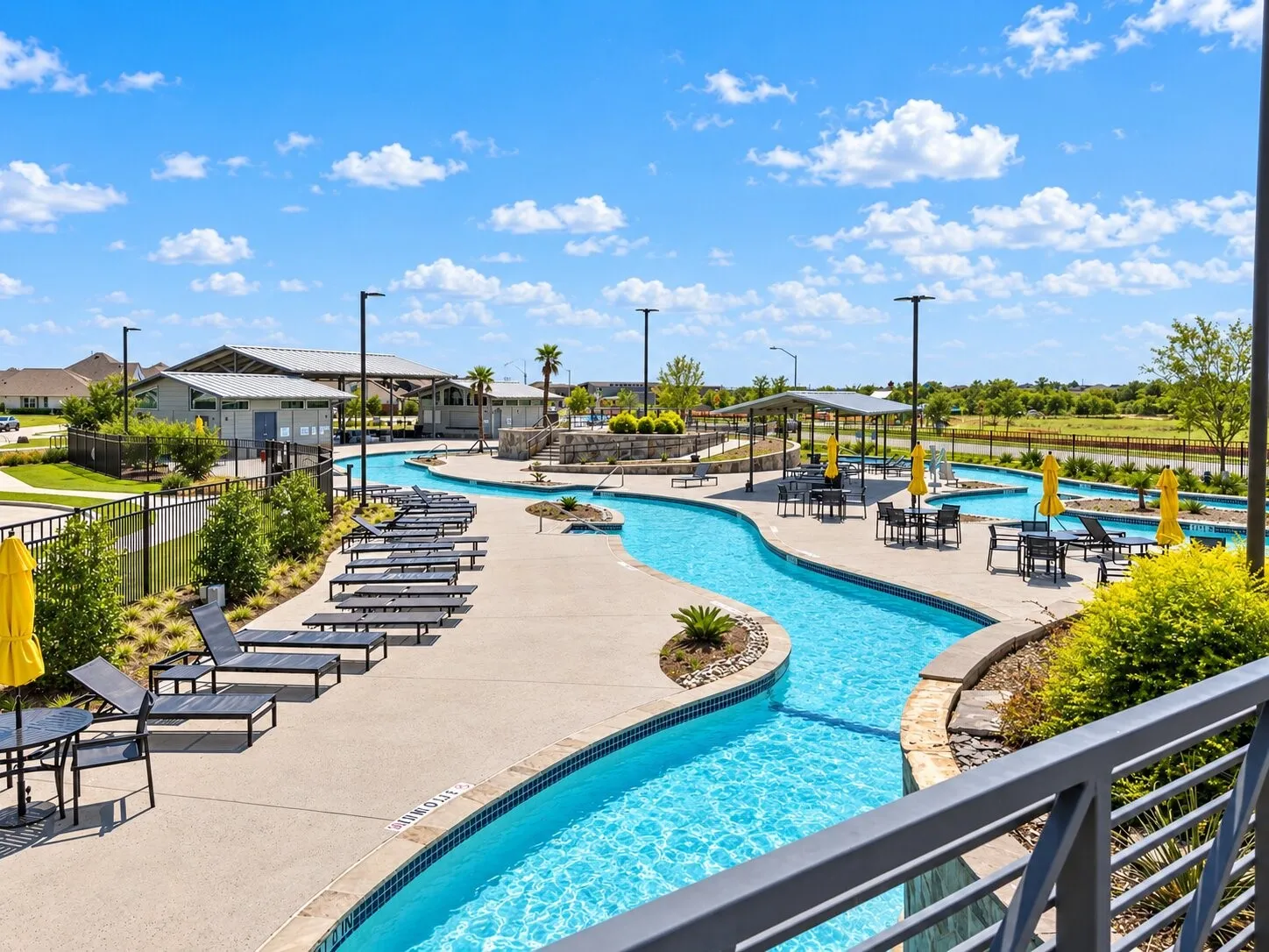 Curving resort-style pool with integrated water features, expansive sundeck with lounge seating, multiple shaded seating areas, and a nearby community building