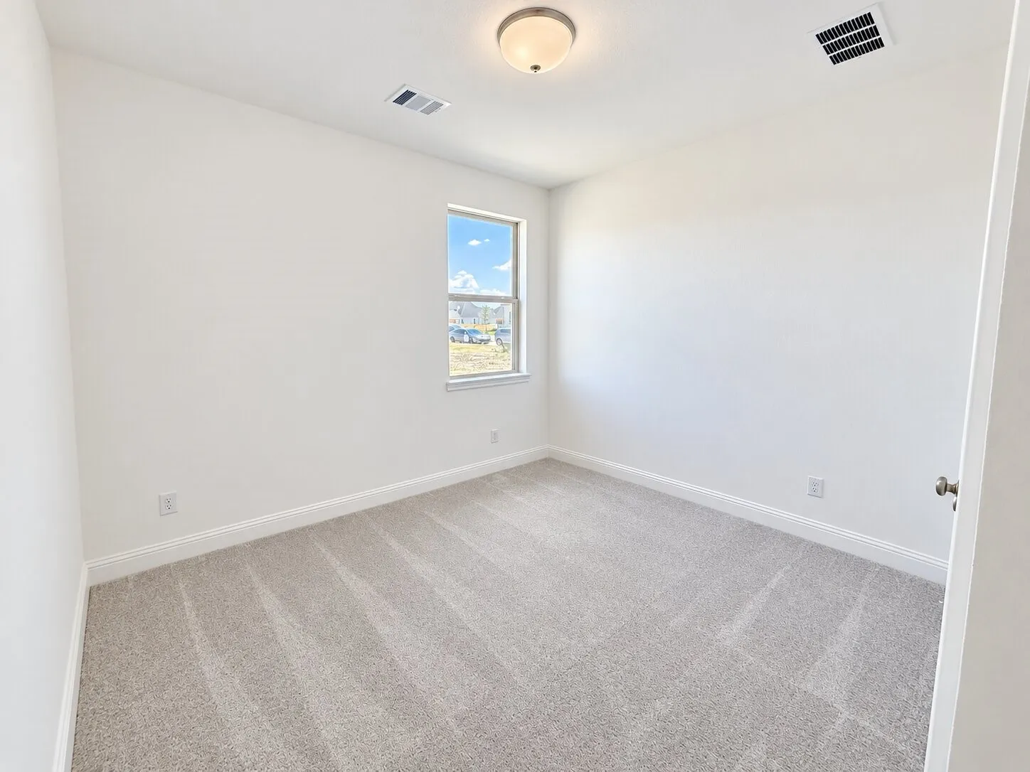 Light gray carpeted flooring paired with white walls, featuring a single window and a semi-flush mount ceiling light