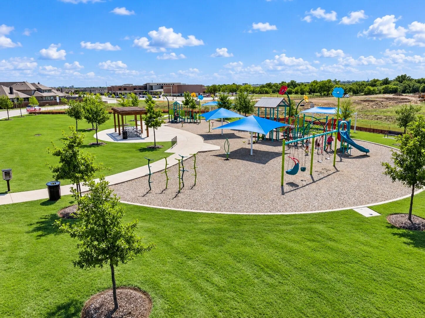 Expansive green space featuring a playground with multiple slides and swings, shaded seating areas, a covered picnic structure, and paved walking paths