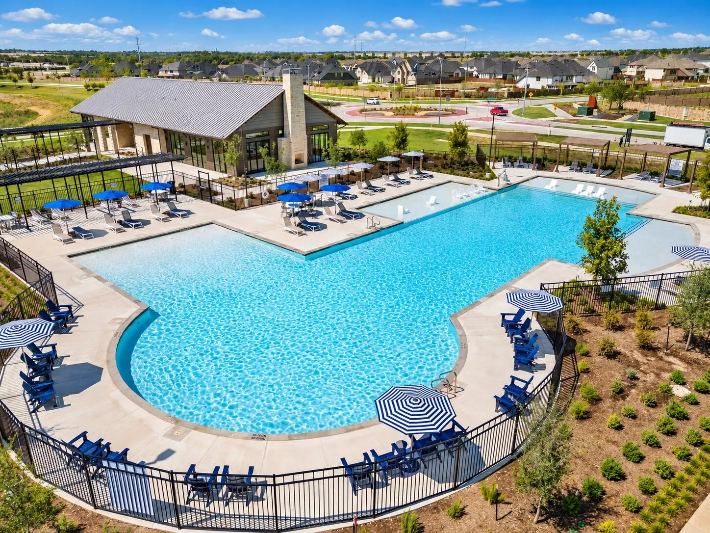 Expansive community pool with multiple swimming areas, surrounding paved deck, and a modern clubhouse featuring a stone chimney