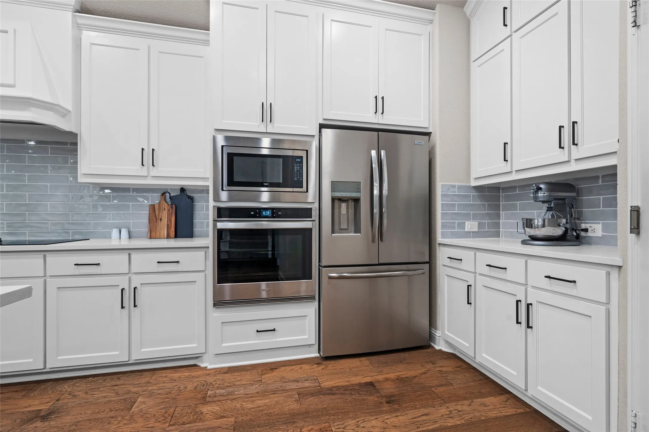 White shaker cabinetry with matte black hardware, stainless steel appliances, tile backsplash, wood-finish flooring, and light-toned countertops
