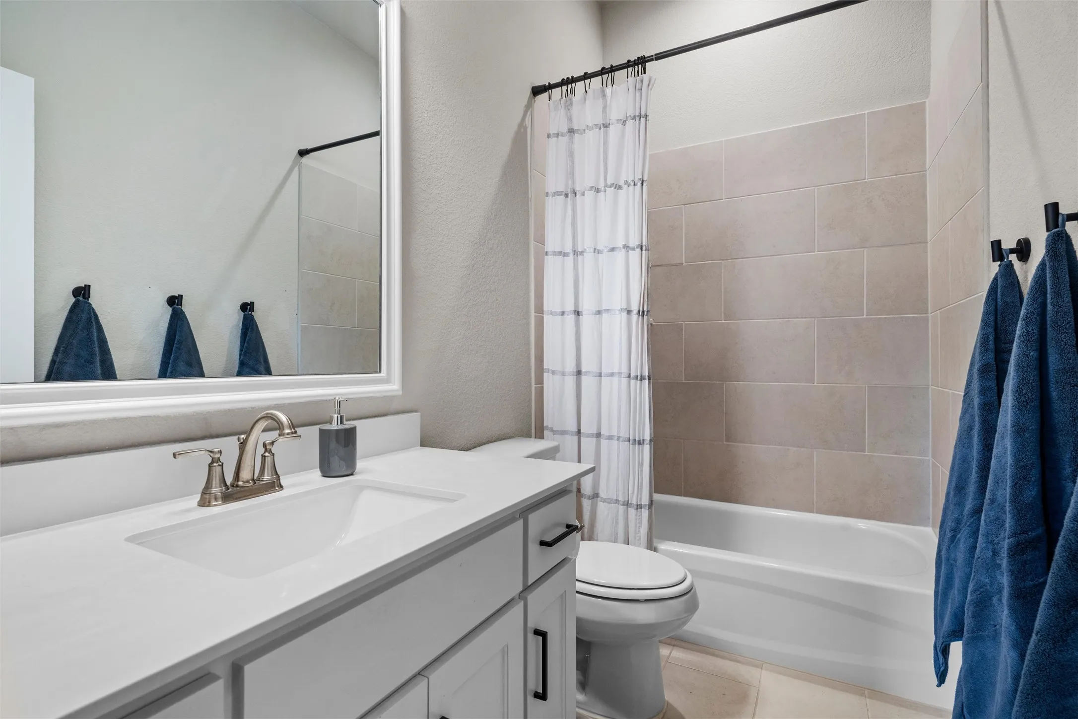 White vanity with integrated sink, brushed nickel faucet, large framed mirror, tiled bathtub surround, and black shower rod