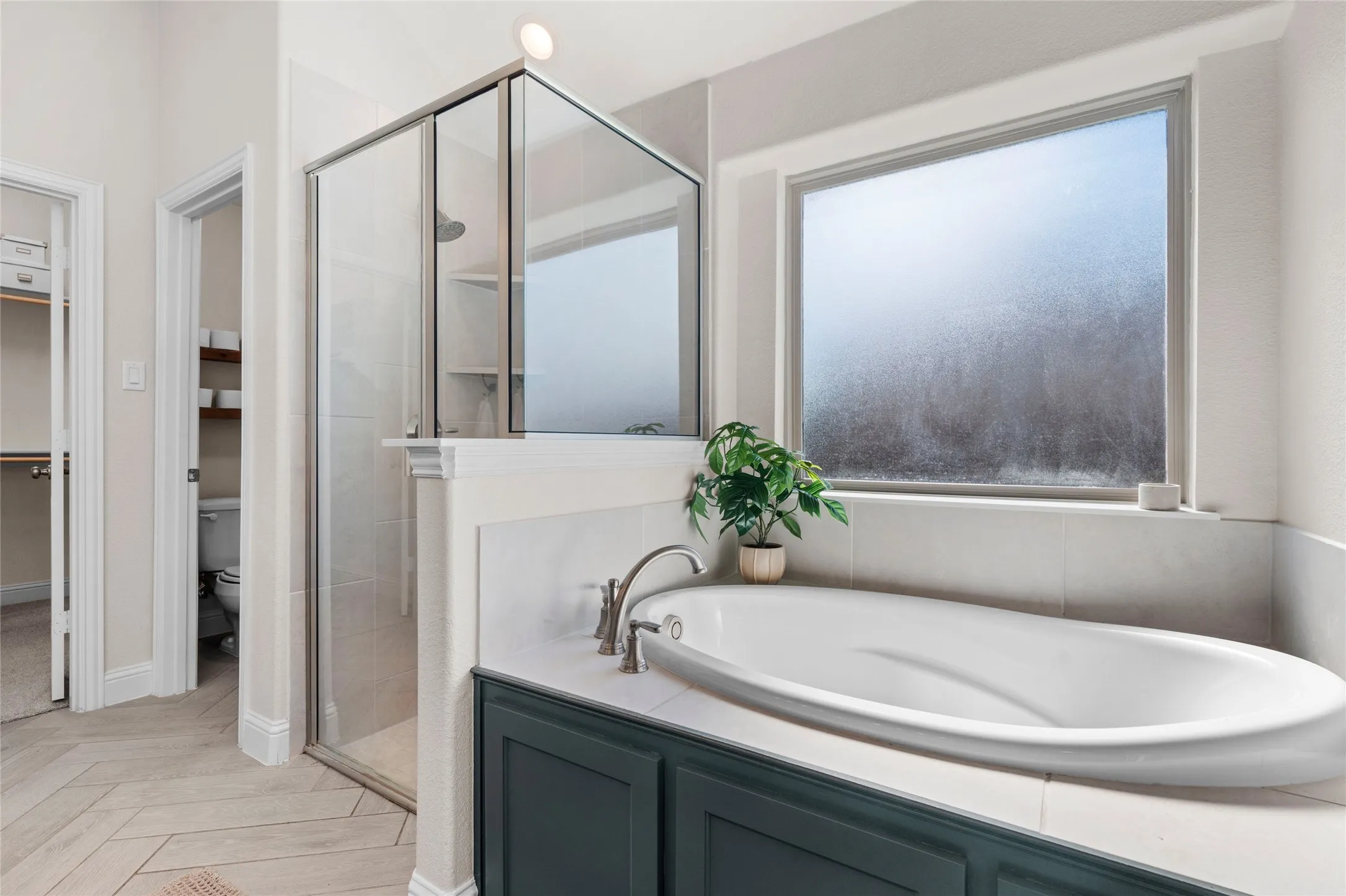 Soaking tub with deck-mounted faucet, walk-in shower with glass enclosure, window with frosted glass, dark-finish cabinetry, and herringbone-pattern wood-finish flooring