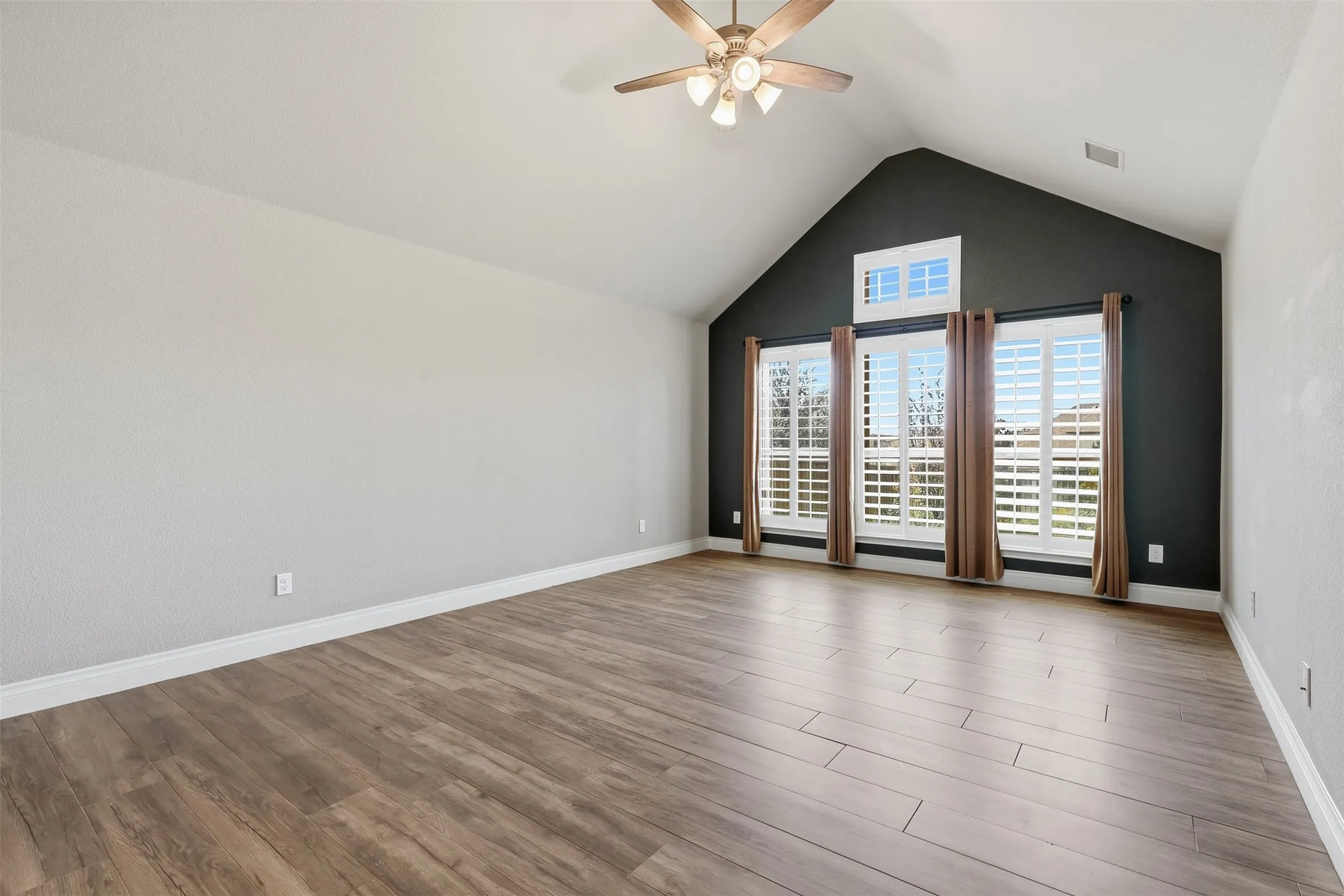 Vaulted ceiling with a ceiling fan, wood-finish flooring, accent wall, and multiple windows with plantation shutters
