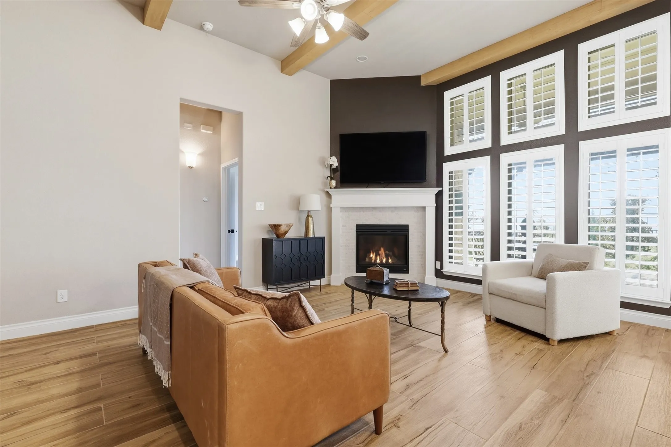 Vaulted ceilings with exposed beams, wood-finish flooring, a contemporary fireplace with a classic mantel, and a multi-pane window wall with plantation shutters