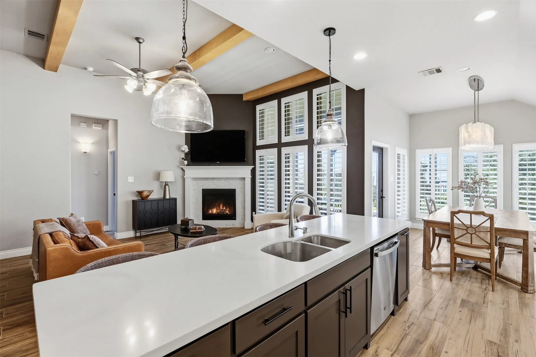 Vaulted ceiling with exposed wood beams, wood-finish flooring, kitchen island with dual sink and stainless steel appliances, corner fireplace with mantel, and multiple windows with plantation shutters