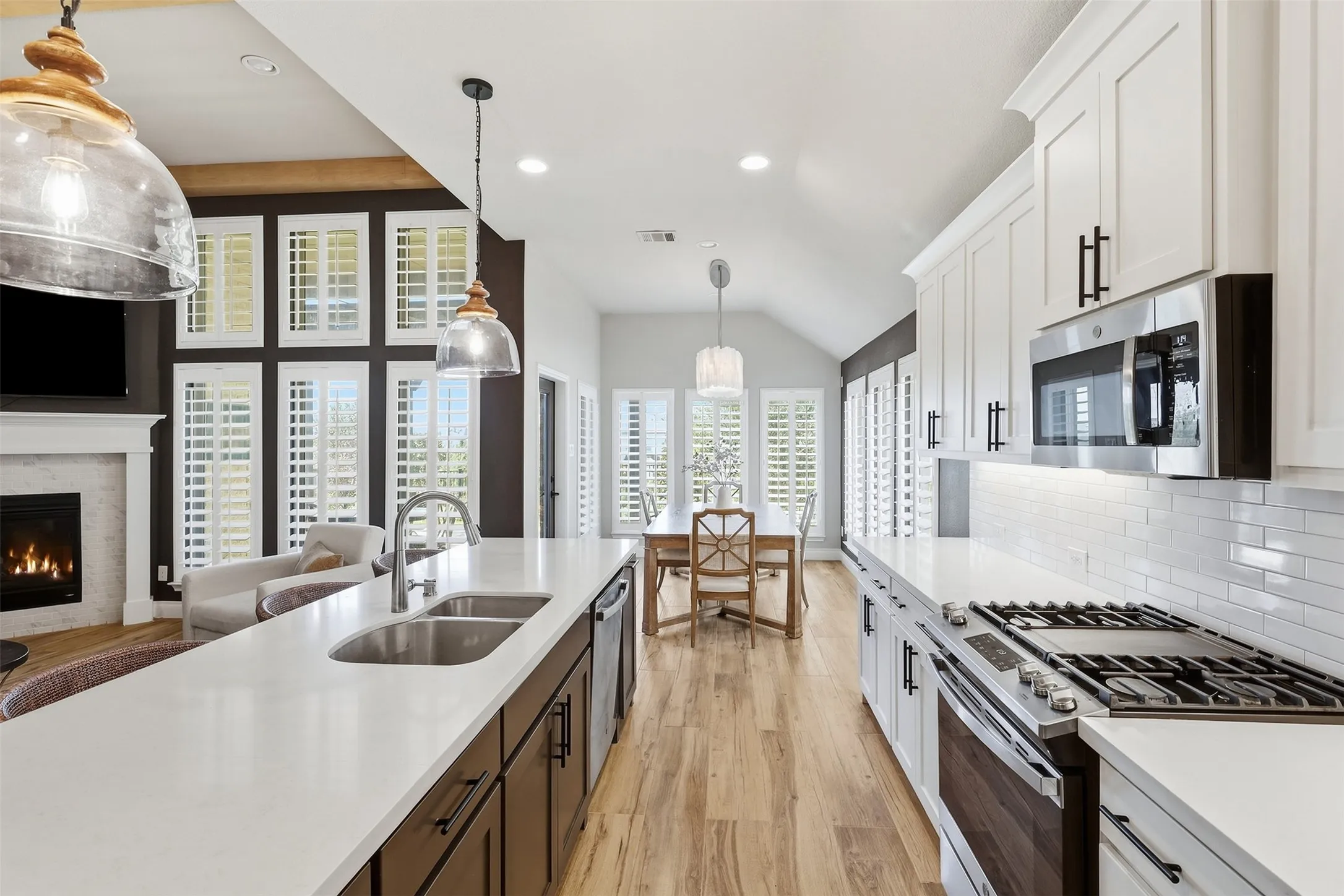 Kitchen island with a double basin sink and a high-arch faucet, white subway tile backsplash, stainless steel appliances, wood-finish flooring, and white plantation shutters