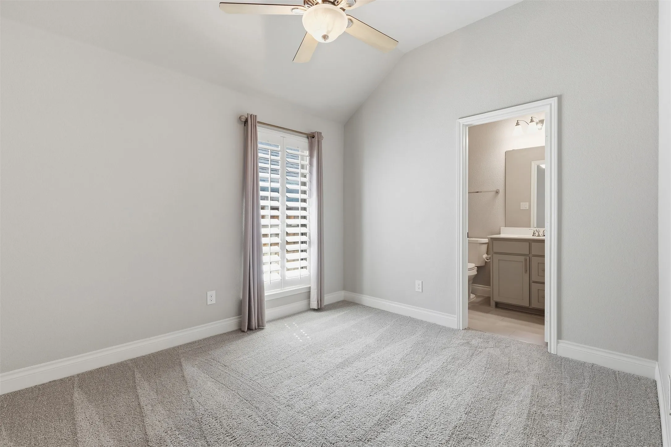 Vaulted ceiling with a ceiling fan, carpet flooring, window with shutters, and interior access to a bathroom