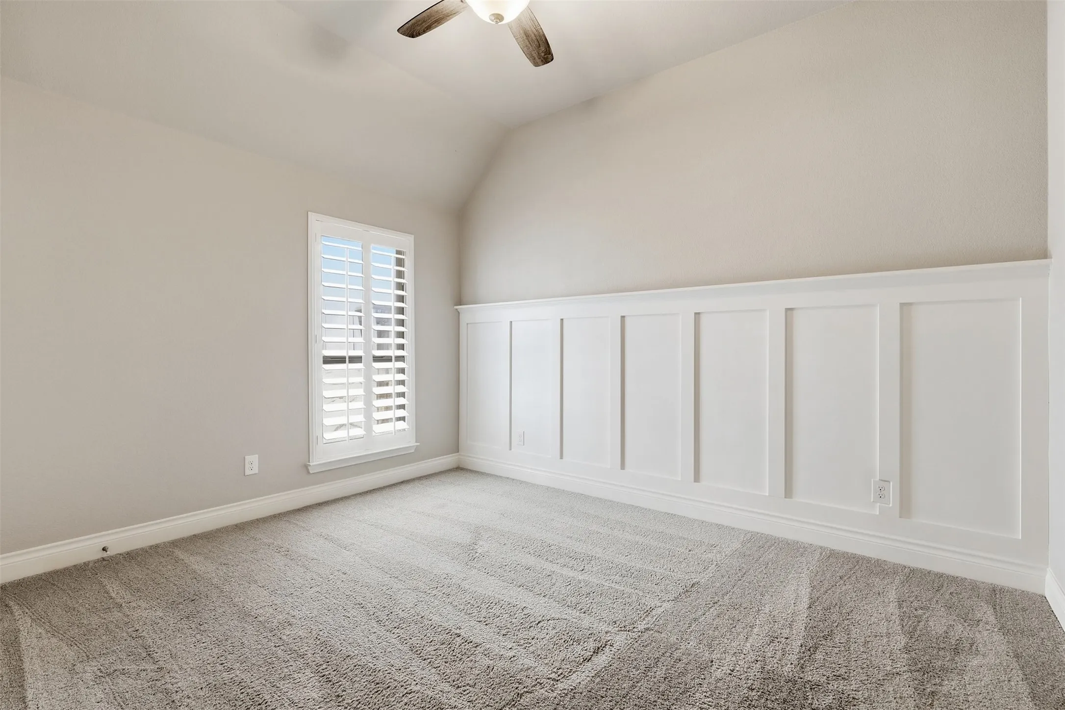 Sloped ceiling, ceiling fan, carpet flooring, window with plantation shutters, and wainscoting detail