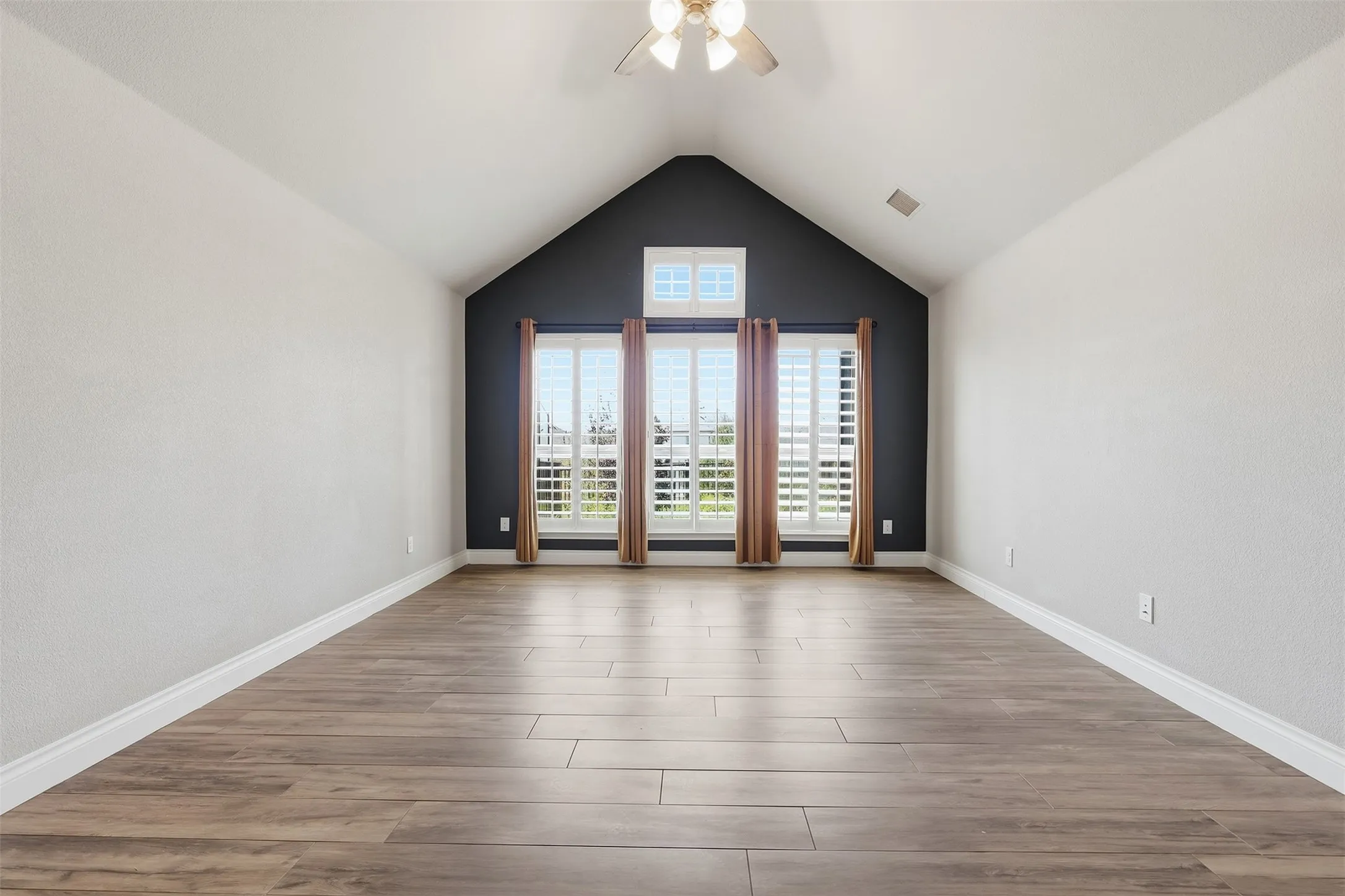 Vaulted ceiling with a ceiling fan, wood-finish flooring, accent wall, and multiple windows with plantation shutters