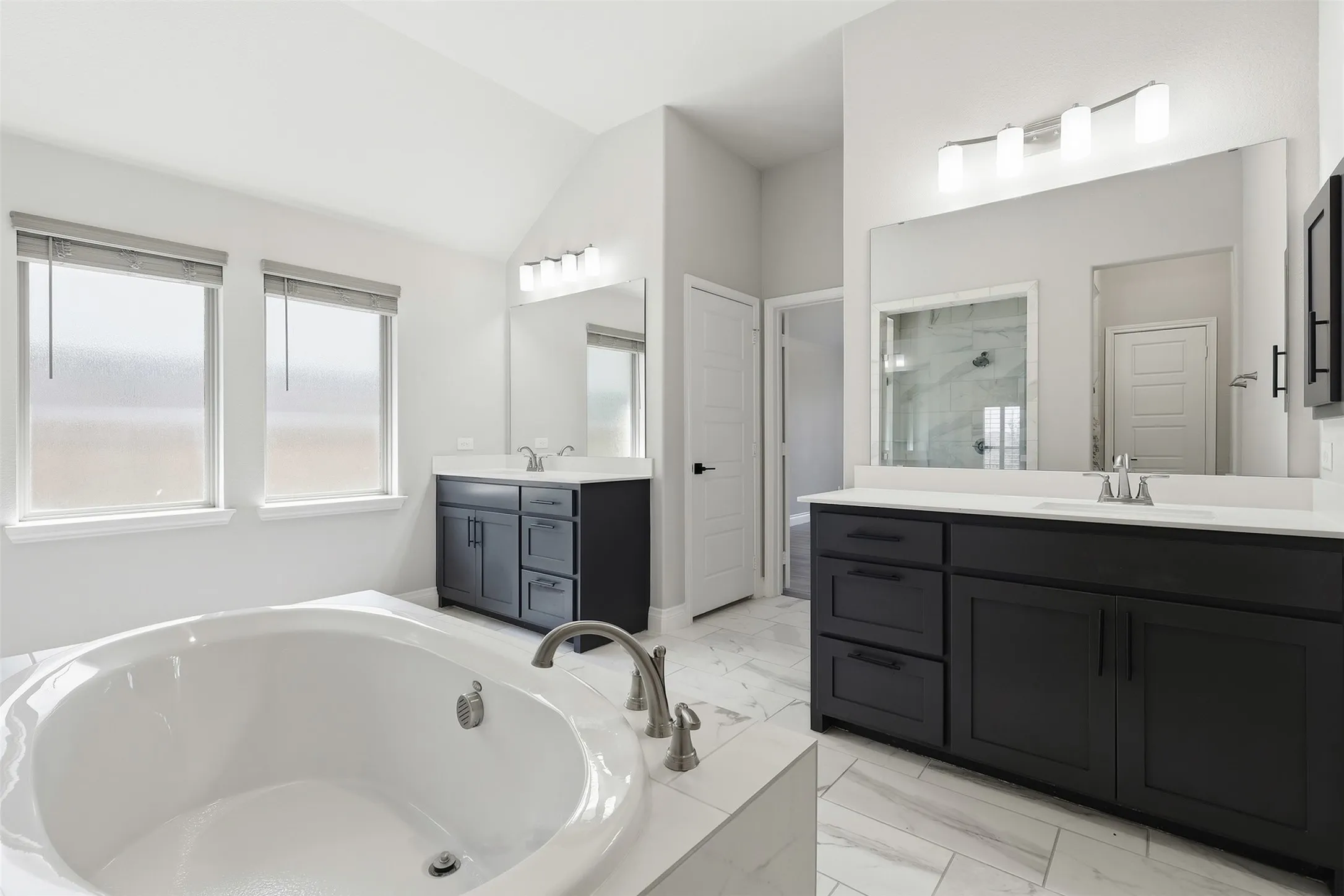 Soaking tub with deck-mounted faucet, dual vanities with dark cabinetry and light countertops, large wall-mounted mirrors, and tiled flooring