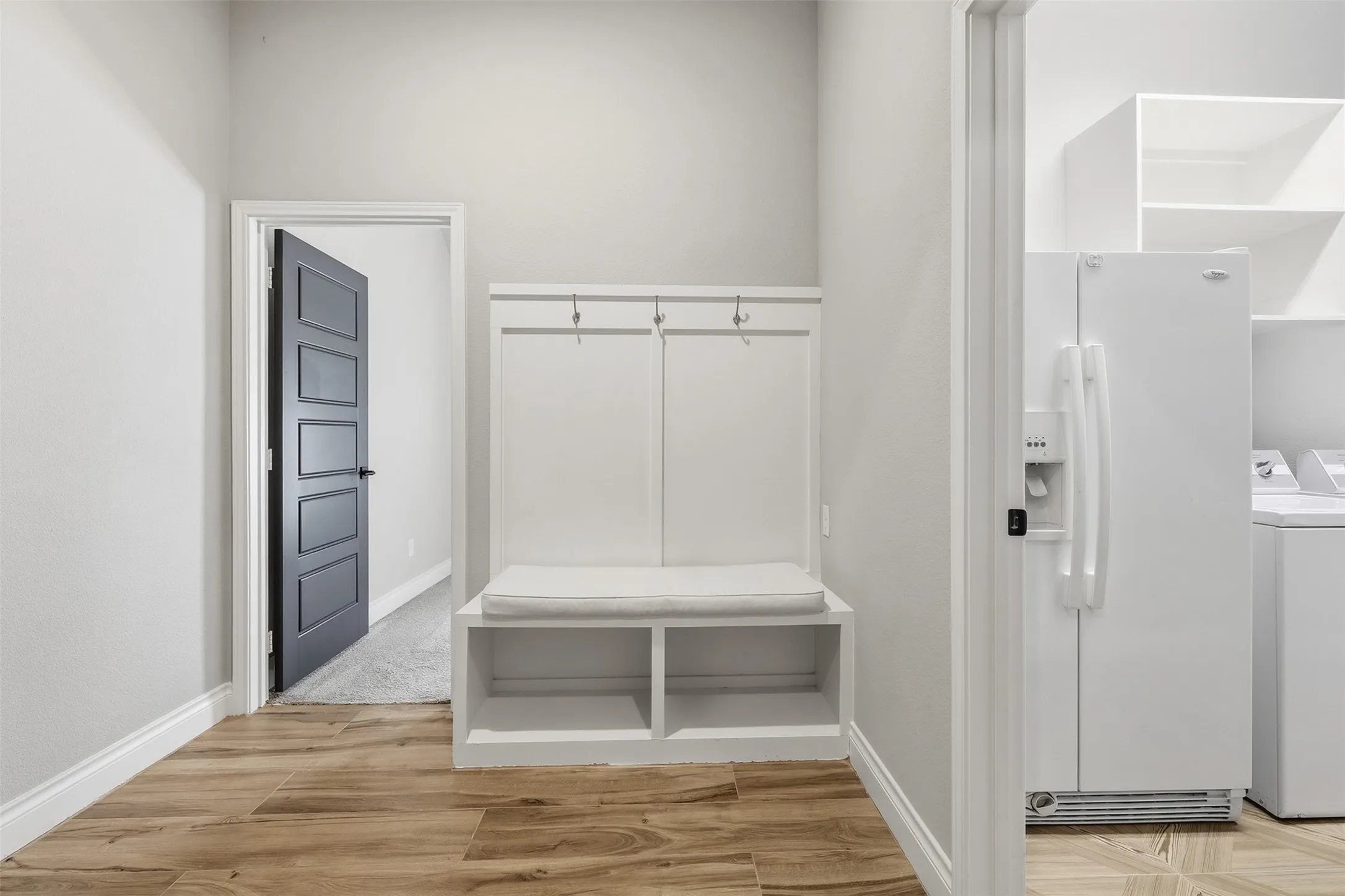 Wood-finish flooring, built-in storage bench with coat hooks, white built-in shelving, white side-by-side refrigerator, and a modern paneled interior door with black hardware