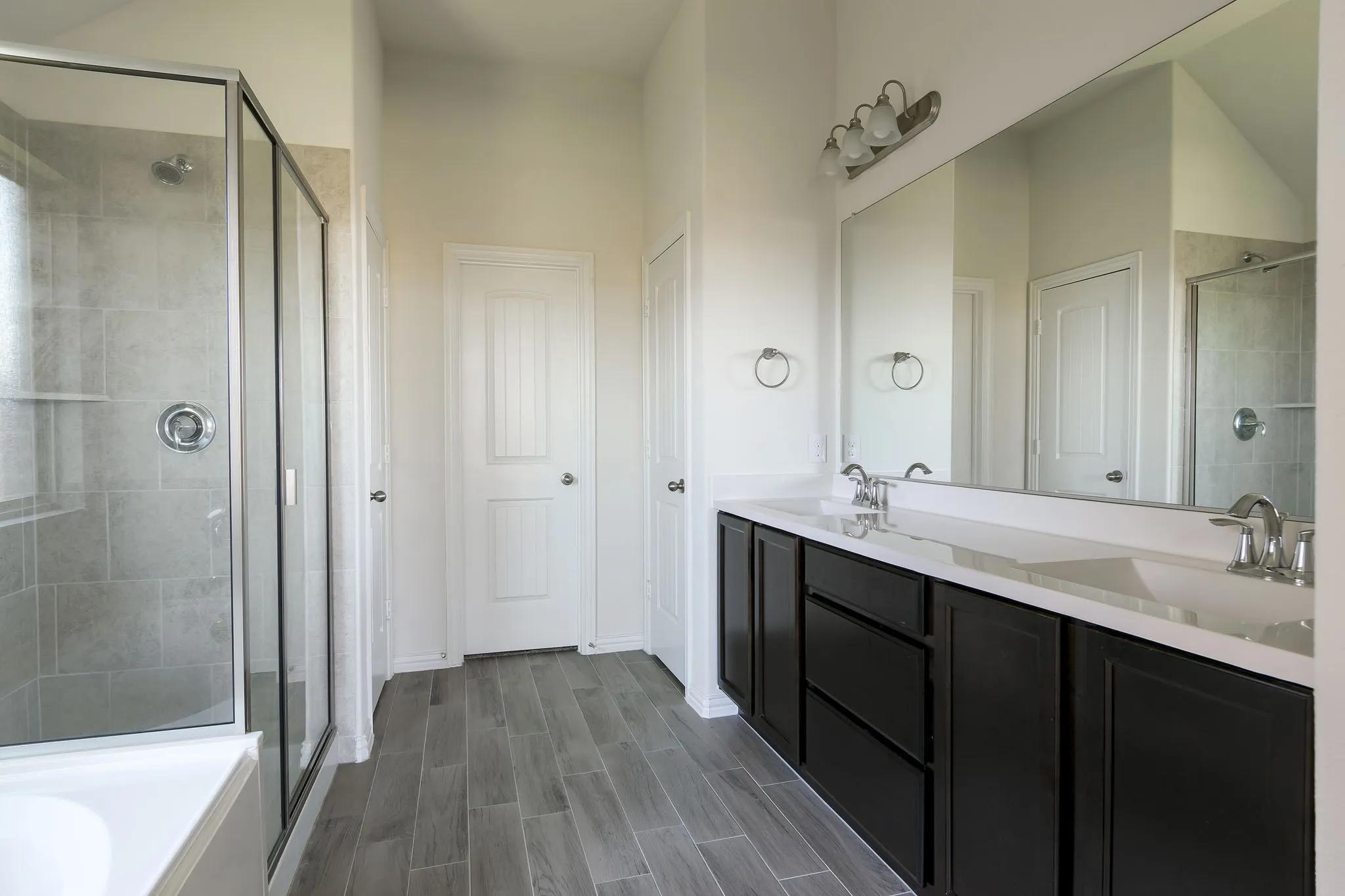 Expansive double vanity with dual sinks, a large mirror, and dark wood-finish cabinetry, complemented by a tiled walk-in shower with a glass enclosure and wood-look flooring